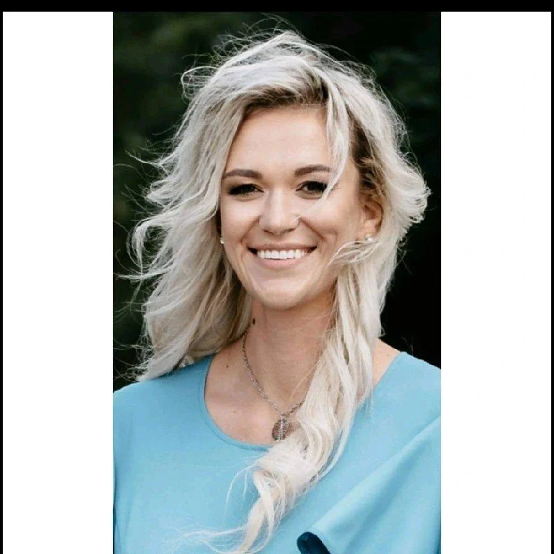 A woman with blonde, wavy hair smiling outdoors, wearing a blue top and a necklace.