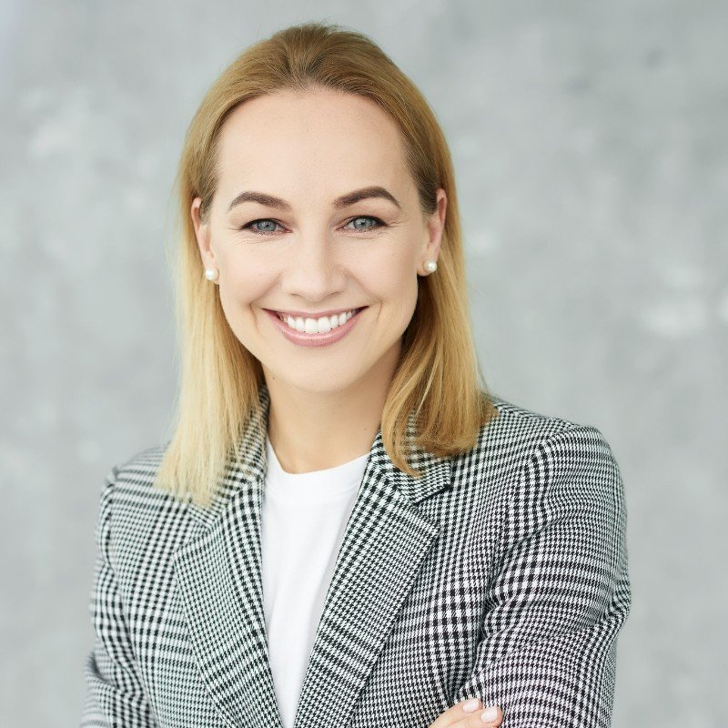 A woman with blonde hair, blue eyes, and fair skin smiling, wearing a checkered blazer and a white shirt against a light gray background.