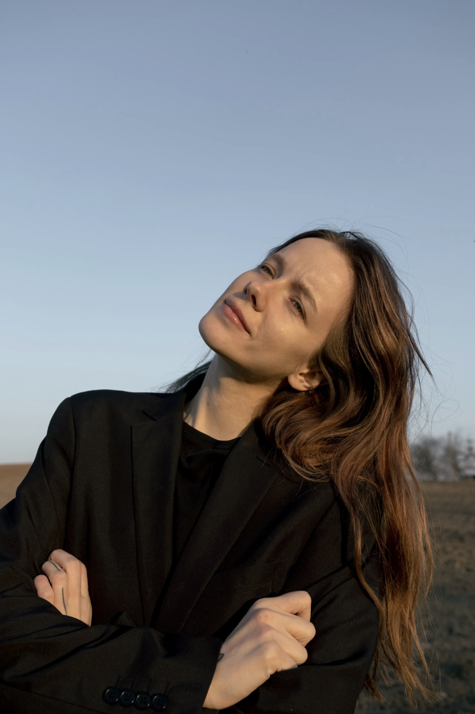 A woman with long, wavy auburn hair and fair skin, wearing a black blazer, stands outdoors with a blue sky in the background. She has a contemplative expression, her head tilted to the side, and her arms crossed over her chest.