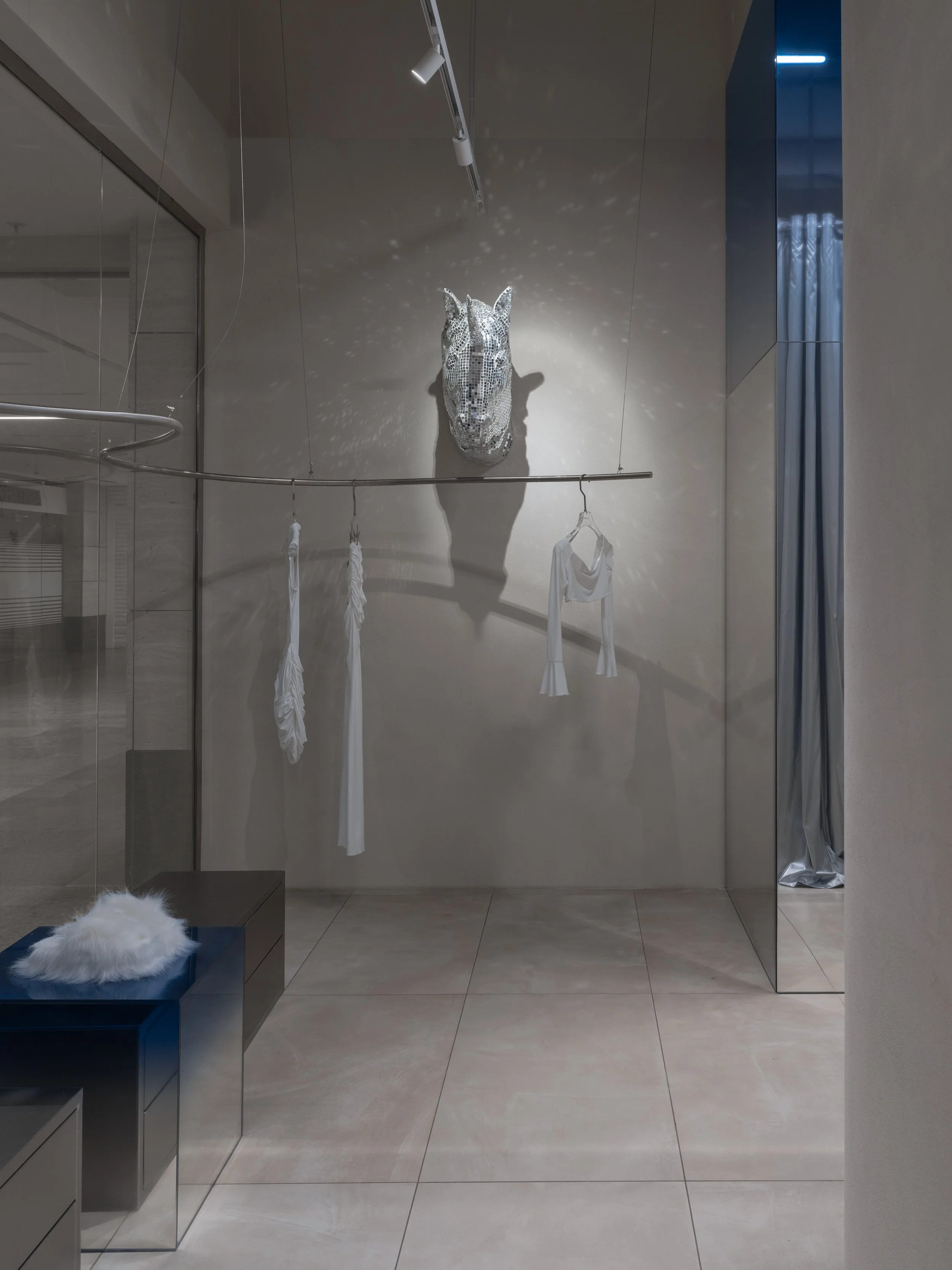 A modern, minimalist interior with a mirrored wall, a silver horse head sculpture casting a shadow, and white clothing hanging on a curved clothesline. A small black table with a white faux fur item is in the corner.