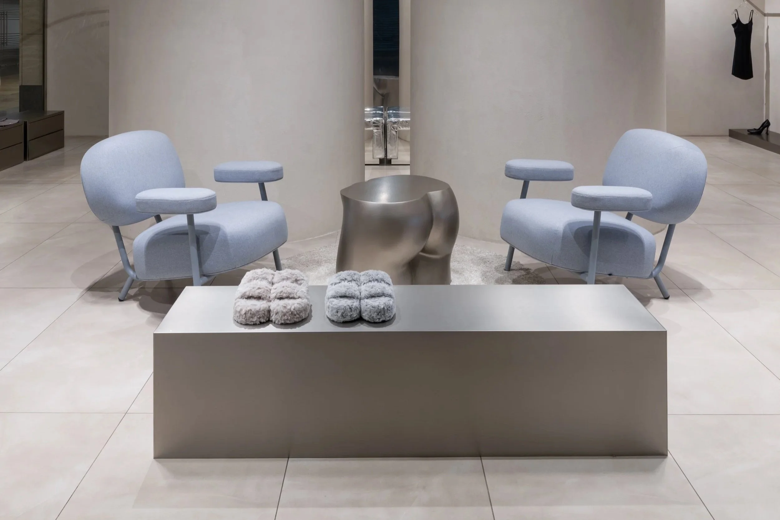Minimalist store display with two light blue armchairs, a metallic sculptural side table, plush slippers, and a clothing rack in background.