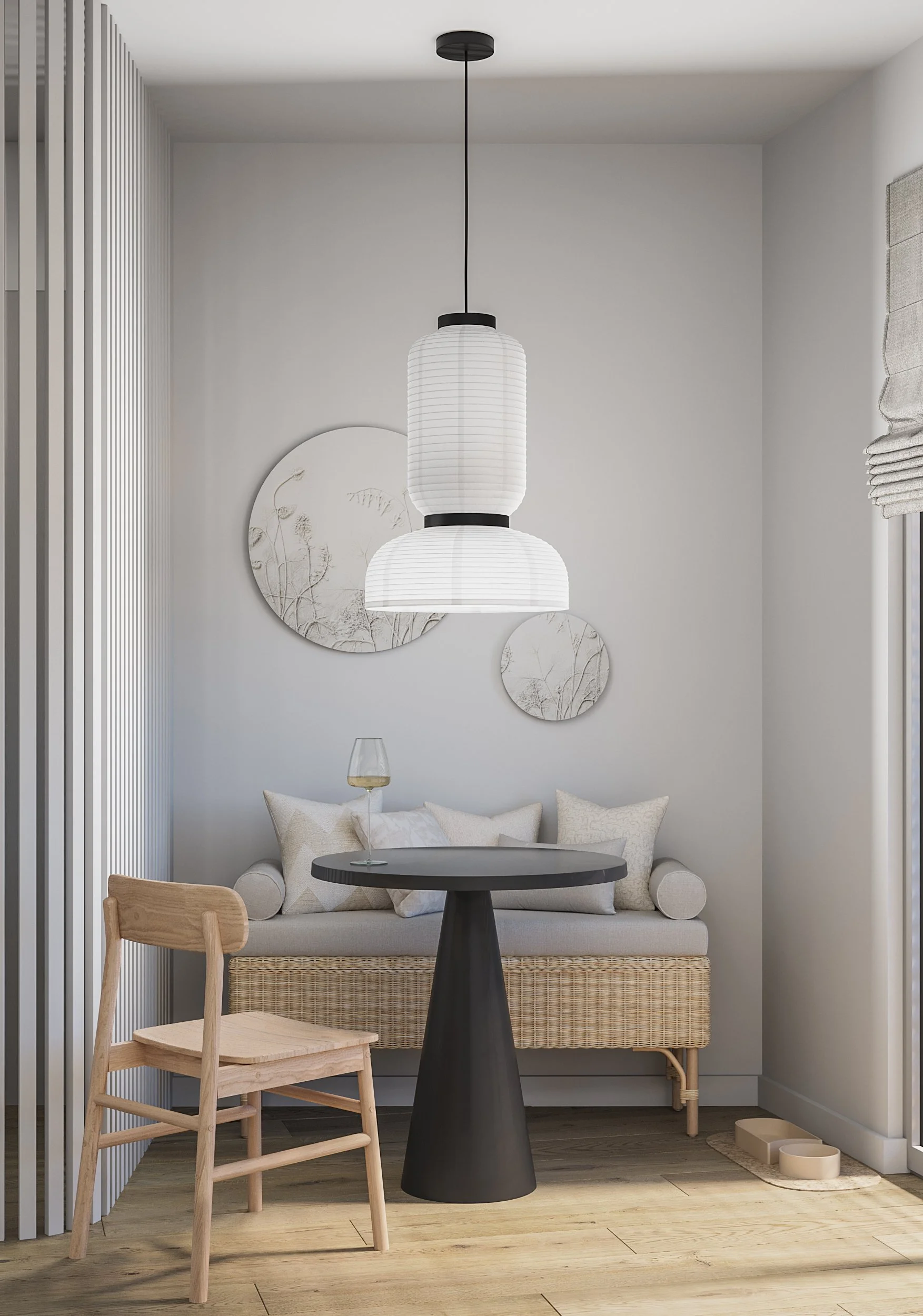 Minimalist interior with a small dining or sitting area featuring a wicker bench with neutral cushions, a black round table, a wooden chair, a hanging white paper lantern light fixture, framed wall art, a glass of wine, and a window with partial blinds.