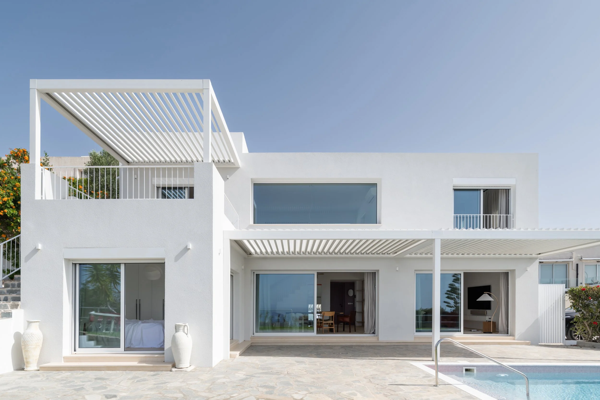 Modern white two-story house with large sliding glass doors, outdoor patio, swimming pool, and minimalistic design under a clear blue sky.