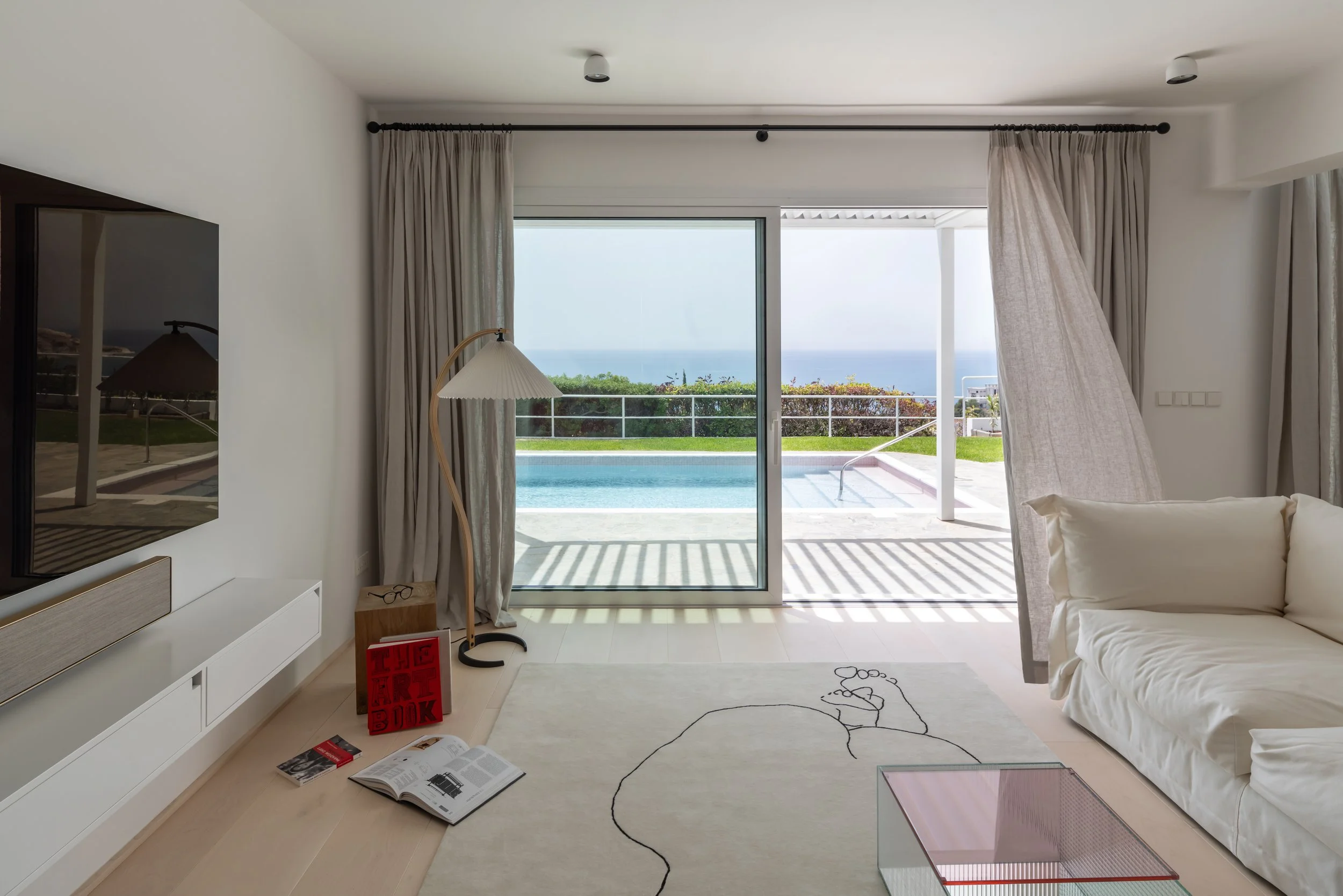 Living room with sliding glass door overlooking a pool and ocean, white sofa, floor lamp, rug with line drawing of a person, books and glasses on the floor, and a TV mounted on the wall.