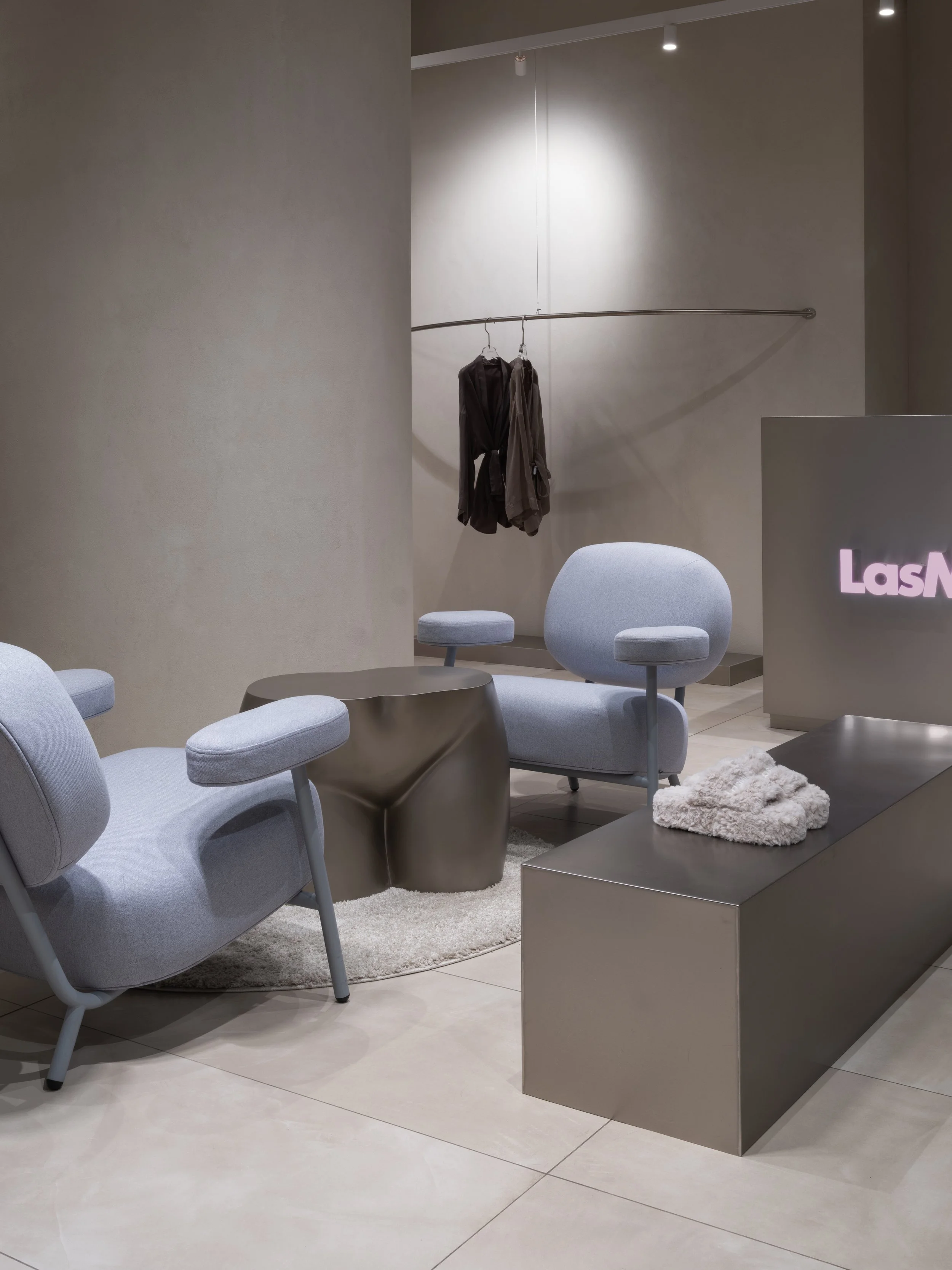 Modern lobby area with two light grey armchairs with curved backs and armrests, a metallic side table with an irregular shape, a small table with a furry cloth, and a coat rack with hanging clothes. There is a reception desk with the partial text 'Las' visible.
