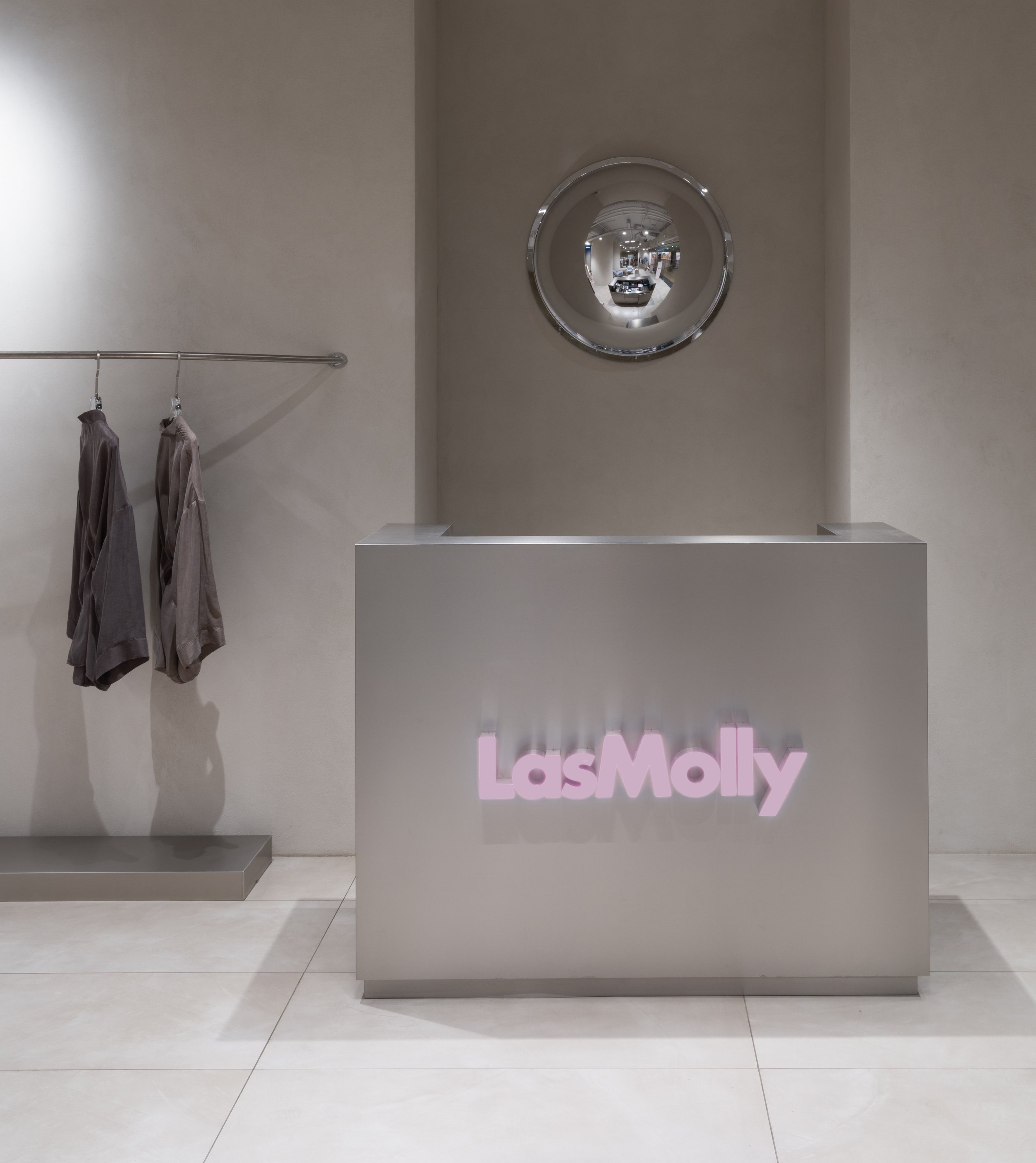 Interior of a boutique with a minimalist design, a reception counter with a glowing 'LasMoly' sign, a mirror on the wall, and a clothing rack with two shirts hanging.