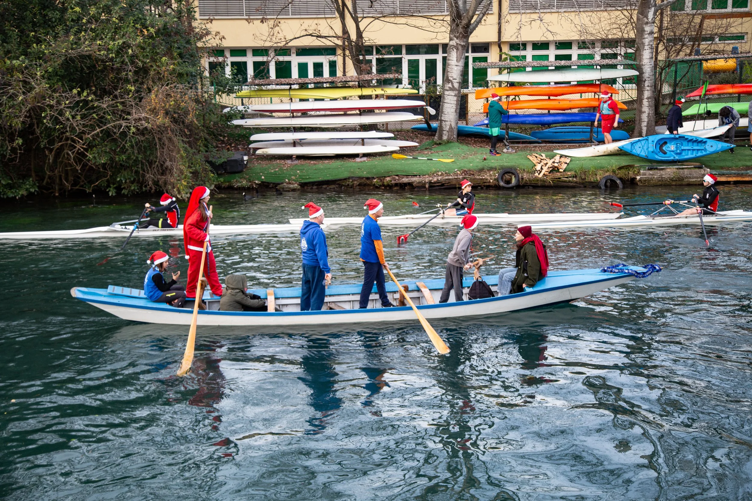 Persone in barca e in kayak con cappelli di Babbo Natale, sugli spaghetti di Natale sul fiume, con kayaks e barche al largo e canoe colorate sulla riva, durante il giorno.