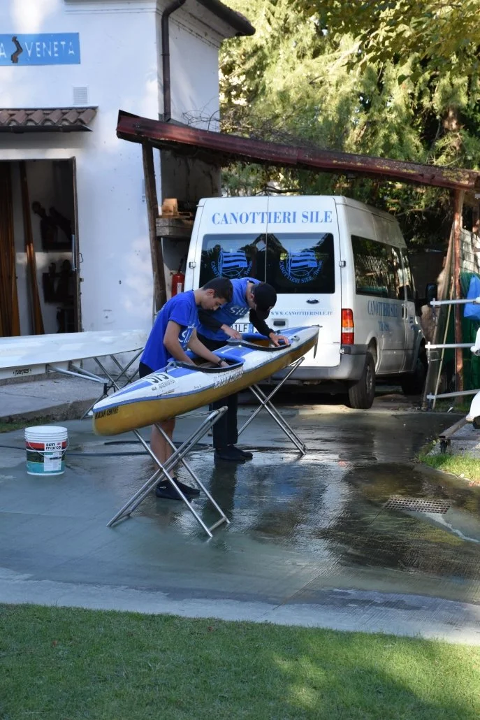 Due giovani sistemano un kayak davanti a un veicolo bianco con scritte in blu sul retro, in un'area all'aperto con alberi e un edificio vicino.