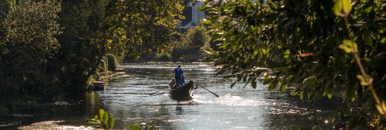 Una barca con due persone che navigano lungo un fiume circondato da alberi verdeggianti.