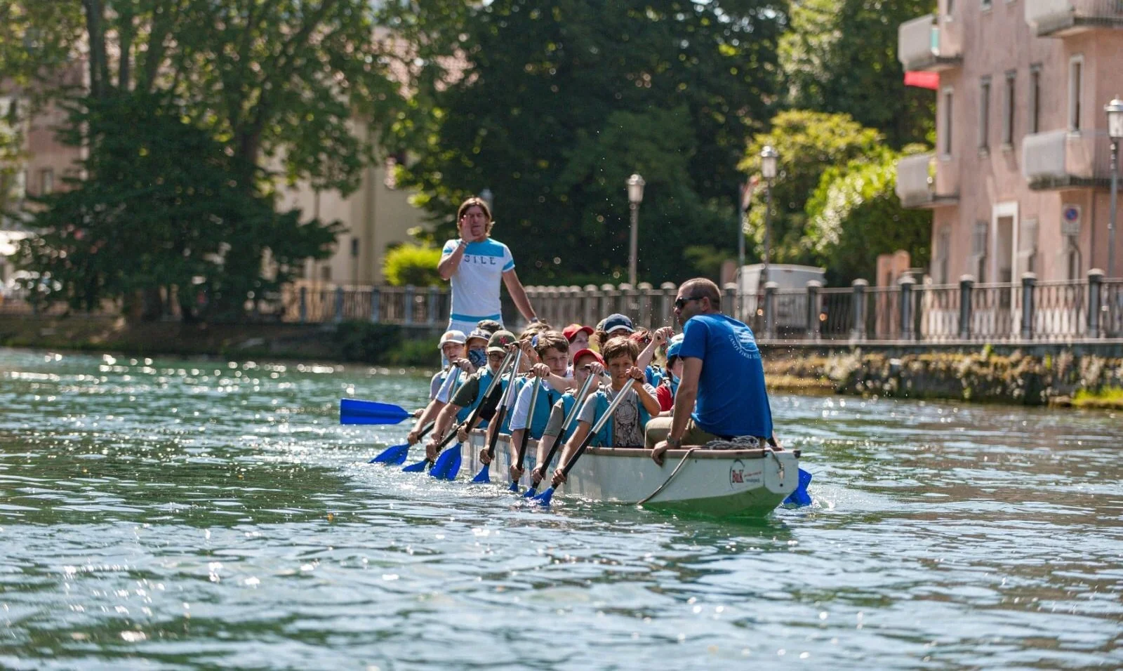 Un gruppo di bambini e un adulto su una barca a remi lungo un fiume, con aria felice, in un ambiente urbano con alberi e edifici.