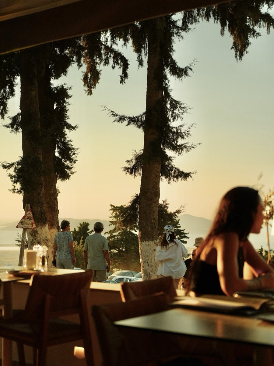 Utsikt över träd och människor som står utomhus i skymningen, sedd från ett inomhuskafé eller restaurang.