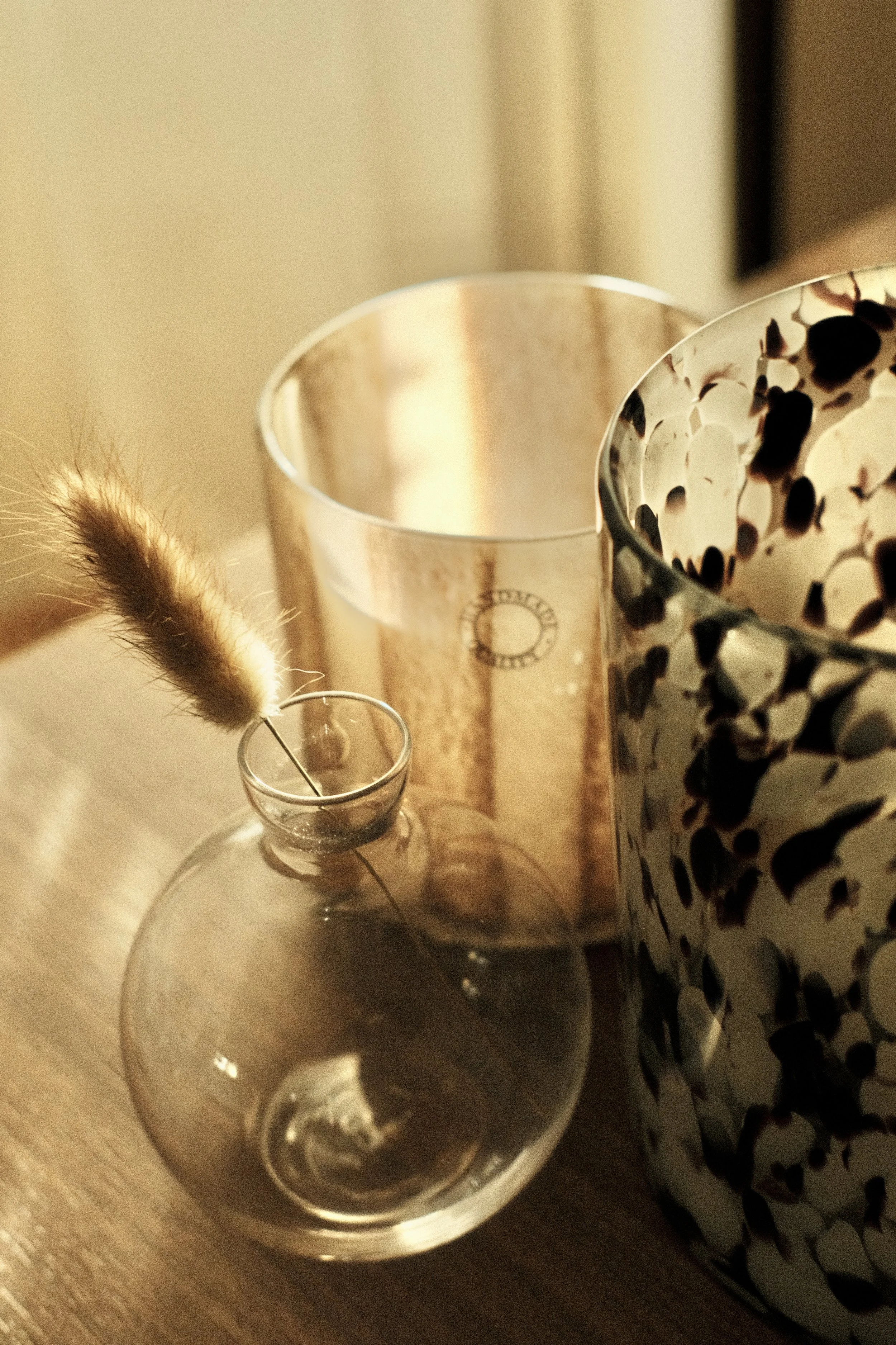 En tom glasflaska med en beige, fjäderliknande dekorationsföremål i koppling till två burkar, en beige och en med svart och vit mönstring, placerade på ett träbord.