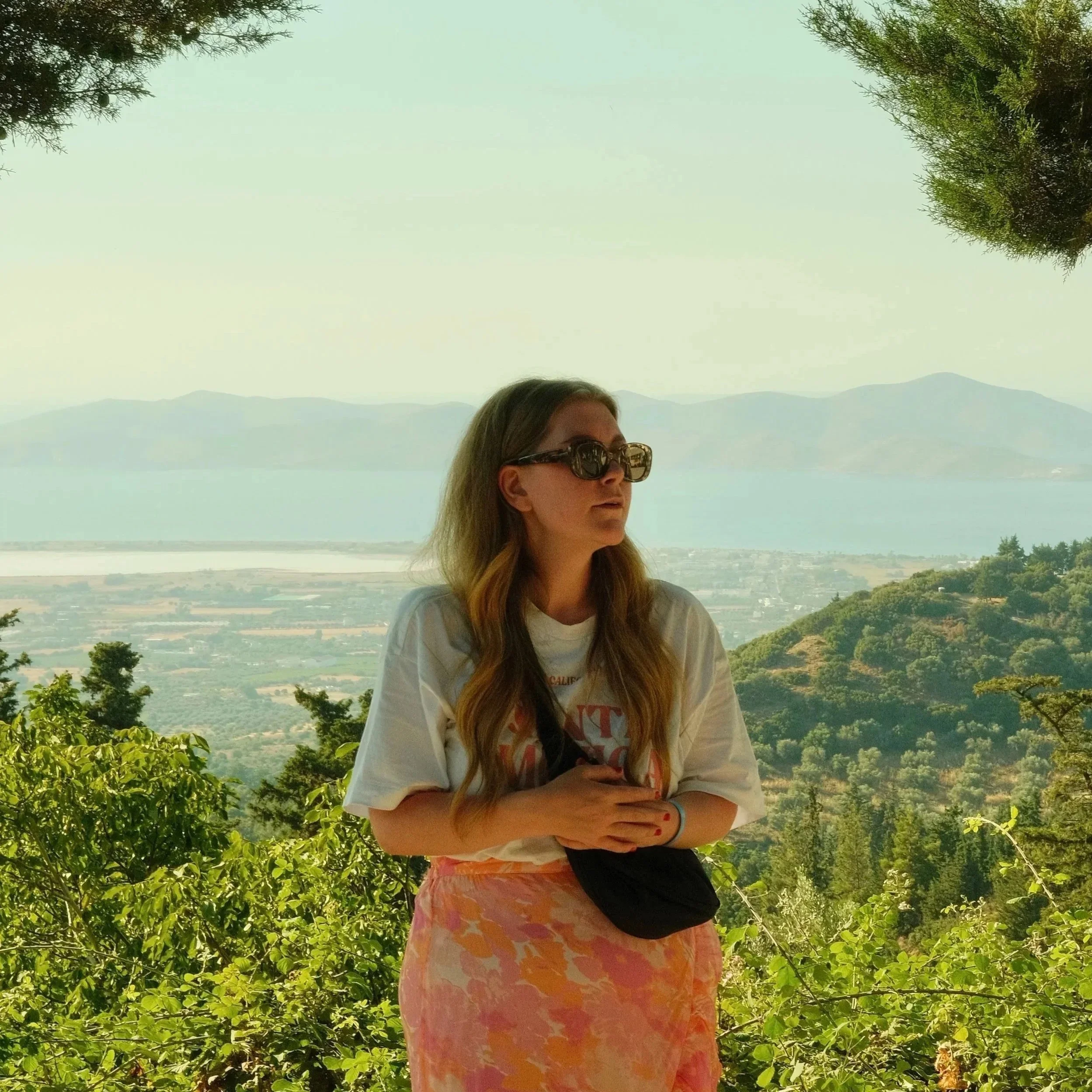 Kvinna med solglasögon står bland gröna buskar med utsikt över träd, vatten och berg i bakgrunden.