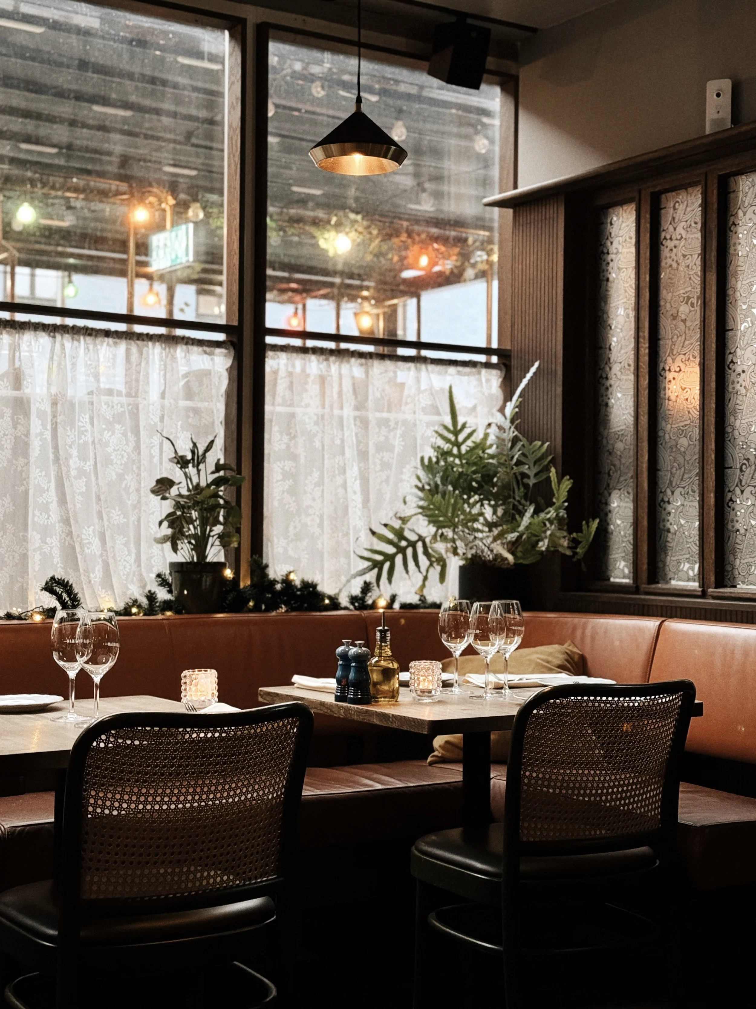 Inbjudande restauranginteriör med bord dukat för middagsgäster, glas, bestick, stearinljus, växter och stora fönster med gardiner och utsikt utomhus.