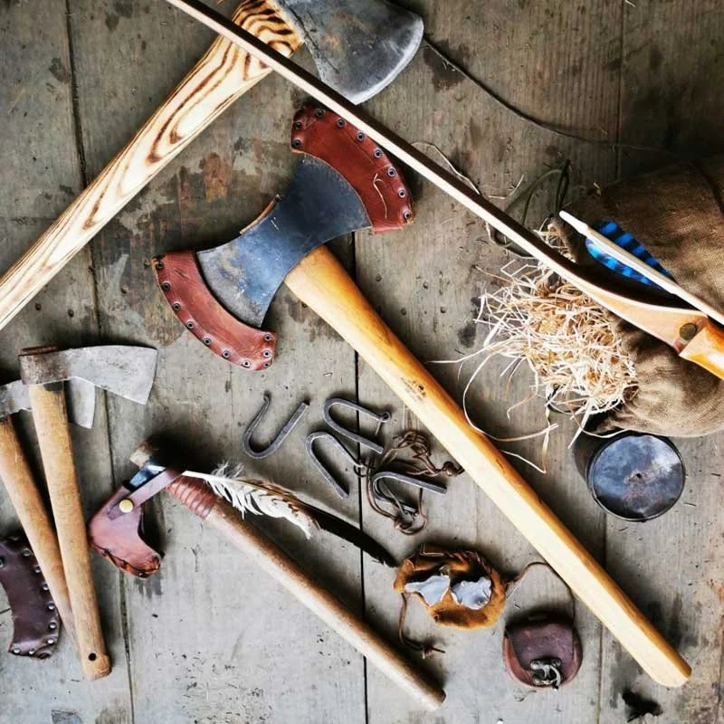 Collection of various hand tools including hatchets, a hammer, a drawknife, metal parts, and wood shavings on a wooden surface.