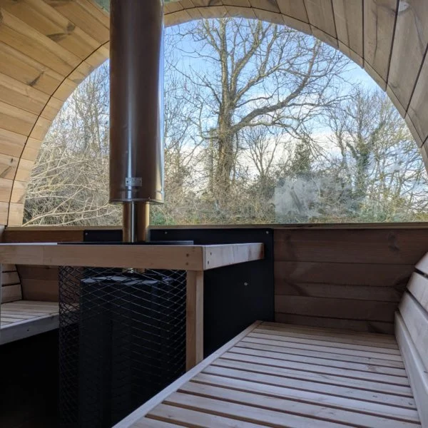 Interior of a wooden sauna with a metal stove and a chimney, large arched window showing trees outside.