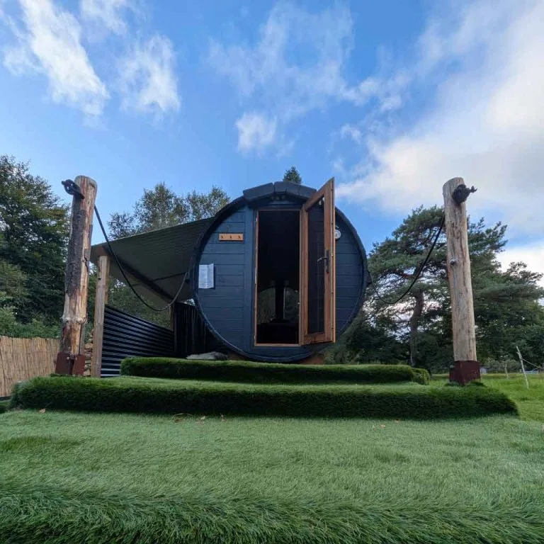 A small black pod-like structure with an open wooden door, elevated on a grassy mound in a park setting, with a blue sky and trees in the background.