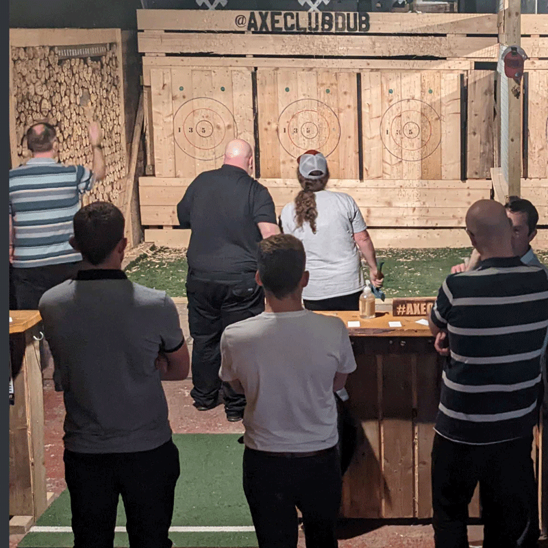 People playing axe throwing at an indoor venue with wooden targets and a sign that says @AXECLUBDUB.