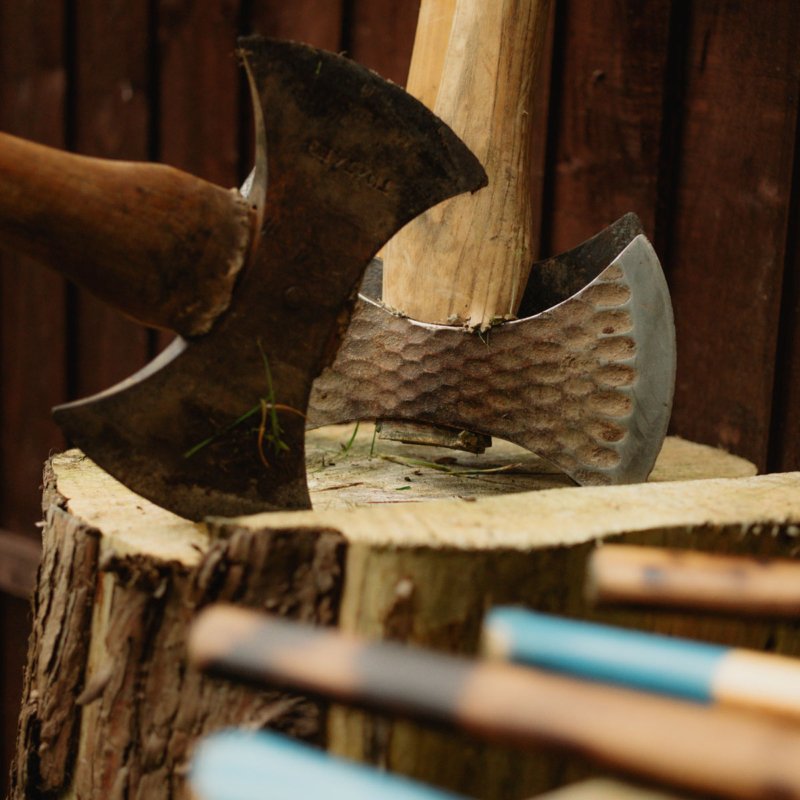 A close-up of an axe embedded in a wooden chopping block with a textured metal head and a wooden handle, set against a dark wooden background.