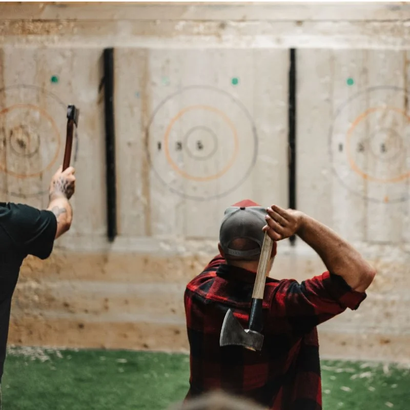 Two men, one wearing a red and black flannel shirt and a baseball cap, are chopping wood in front of an axe throwing target wall with three painted circles. The second man is holding an axe in the air, preparing to throw.