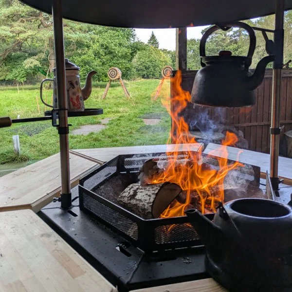 Close-up of a campfire with a metal kettle hanging above the flames on a grill setup outdoors surrounded by green trees and grass.
