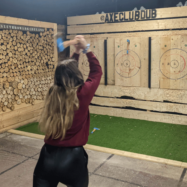 A woman with long hair wearing a maroon hoodie and black pants swinging an axe at an axe throwing target in an indoor axe throwing alley.