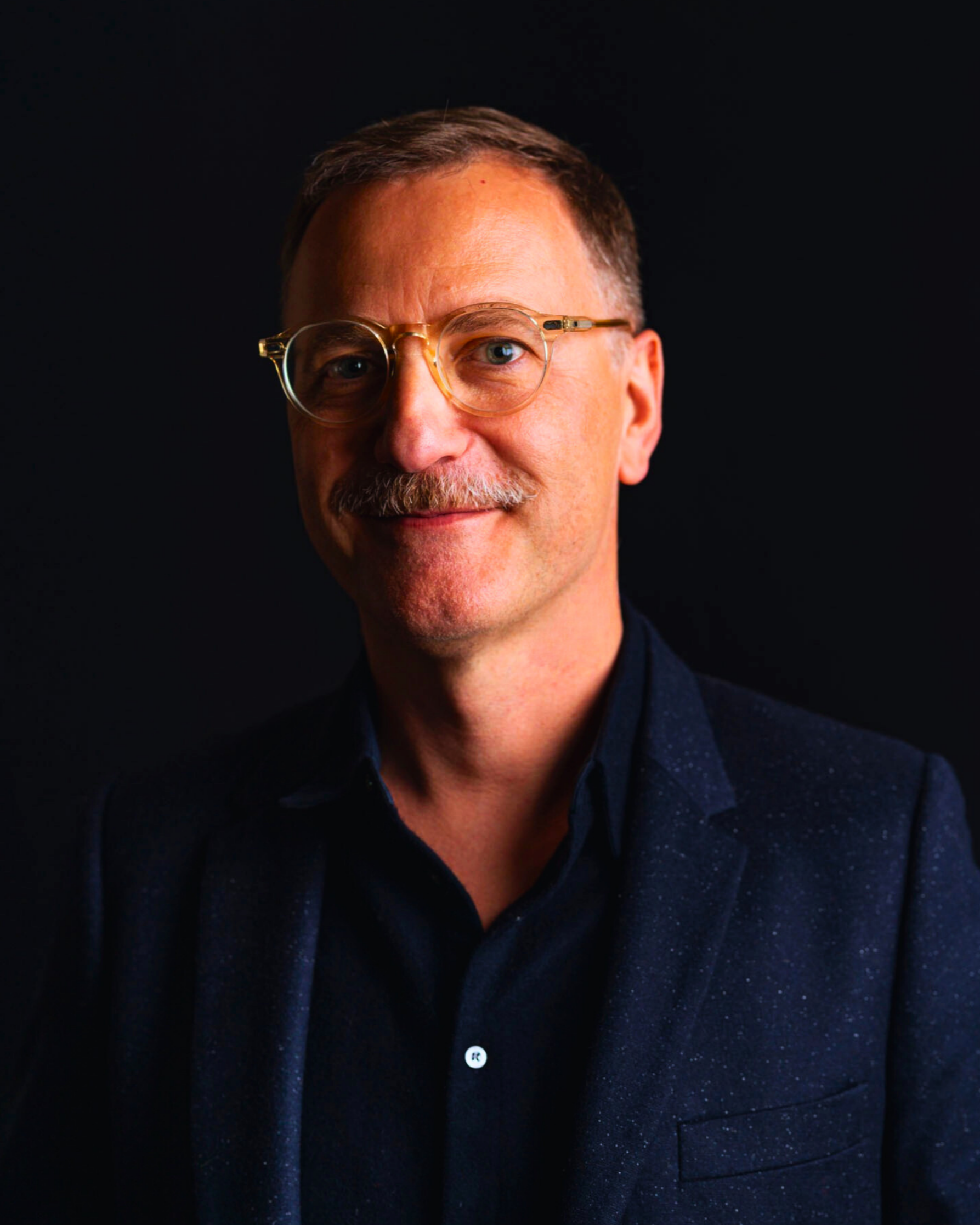 Close-up portrait of a man with glasses, a mustache, and short brown hair, wearing a dark, possibly navy, dotted shirt, against a black background.