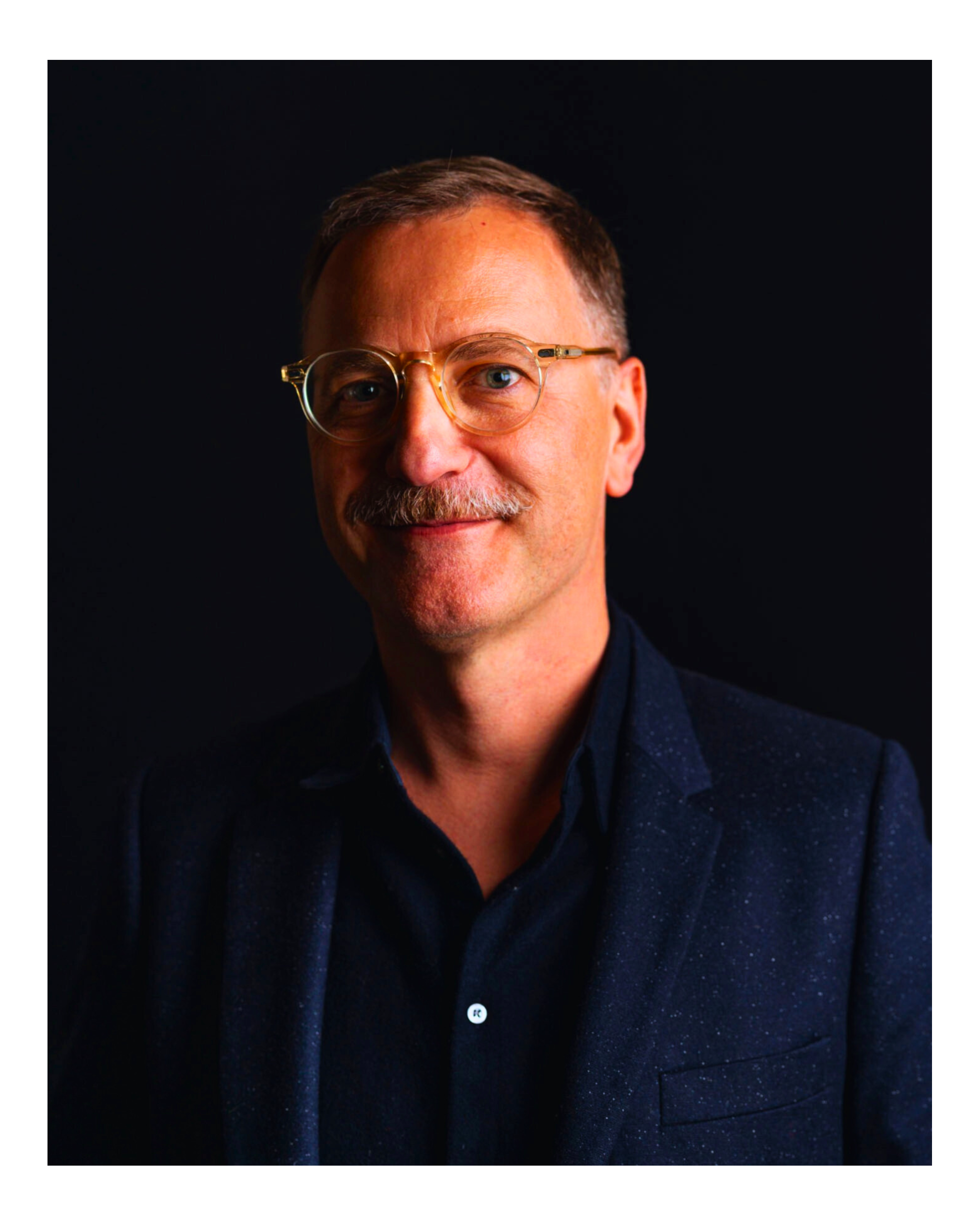 Close-up portrait of a middle-aged man with short brown hair, wearing gold-rimmed glasses, a dark blue speckled blazer, and a black shirt, against a black background.