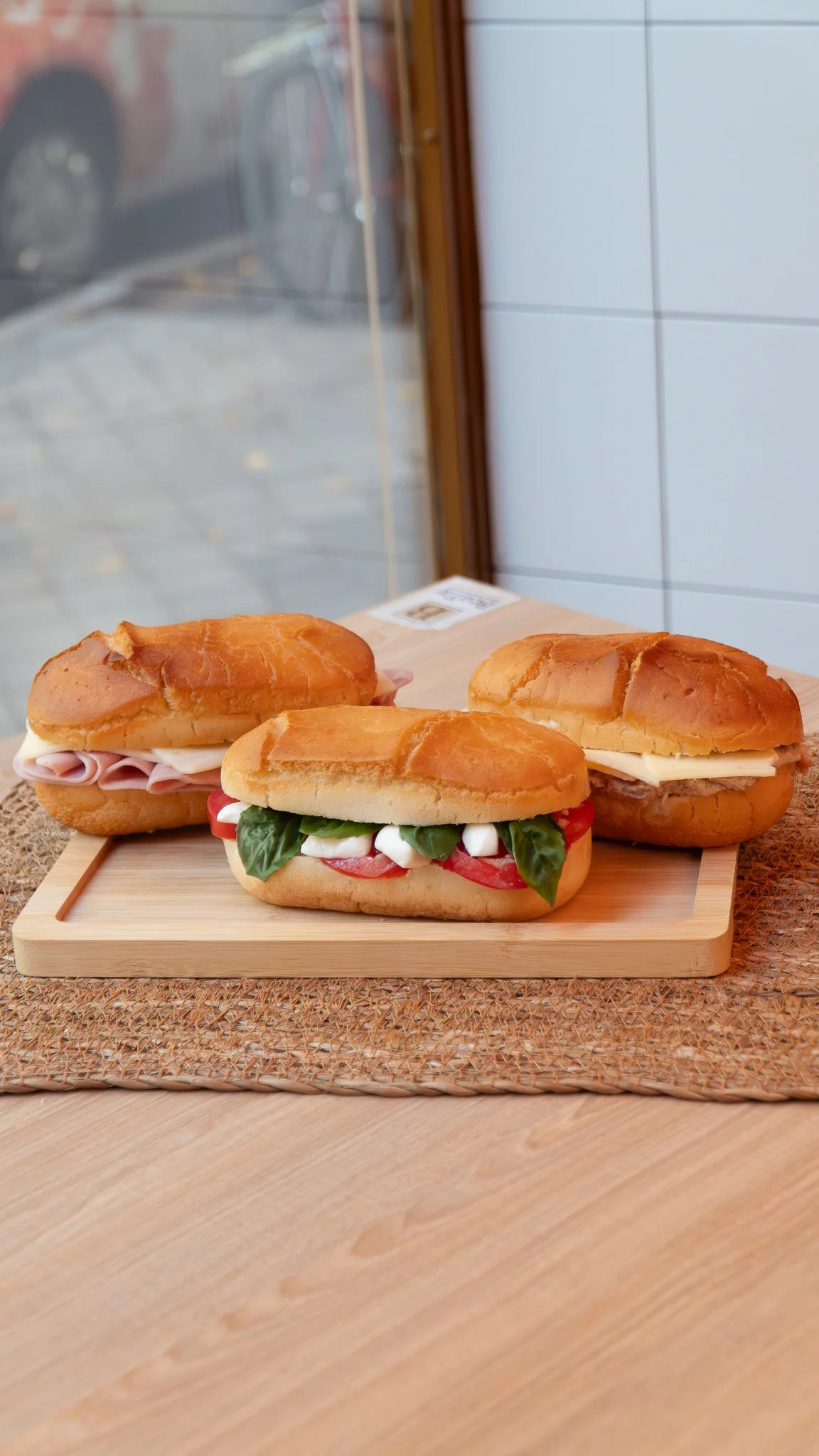 Tres sándwiches en un plato de madera sobre una mesa, con una ventana al fondo
