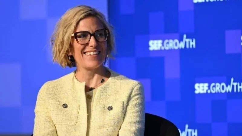 A woman with blonde hair and glasses smiling, wearing a light-colored jacket, sitting in front of a blue background with white text that says 'SEE. GROWTH'.
