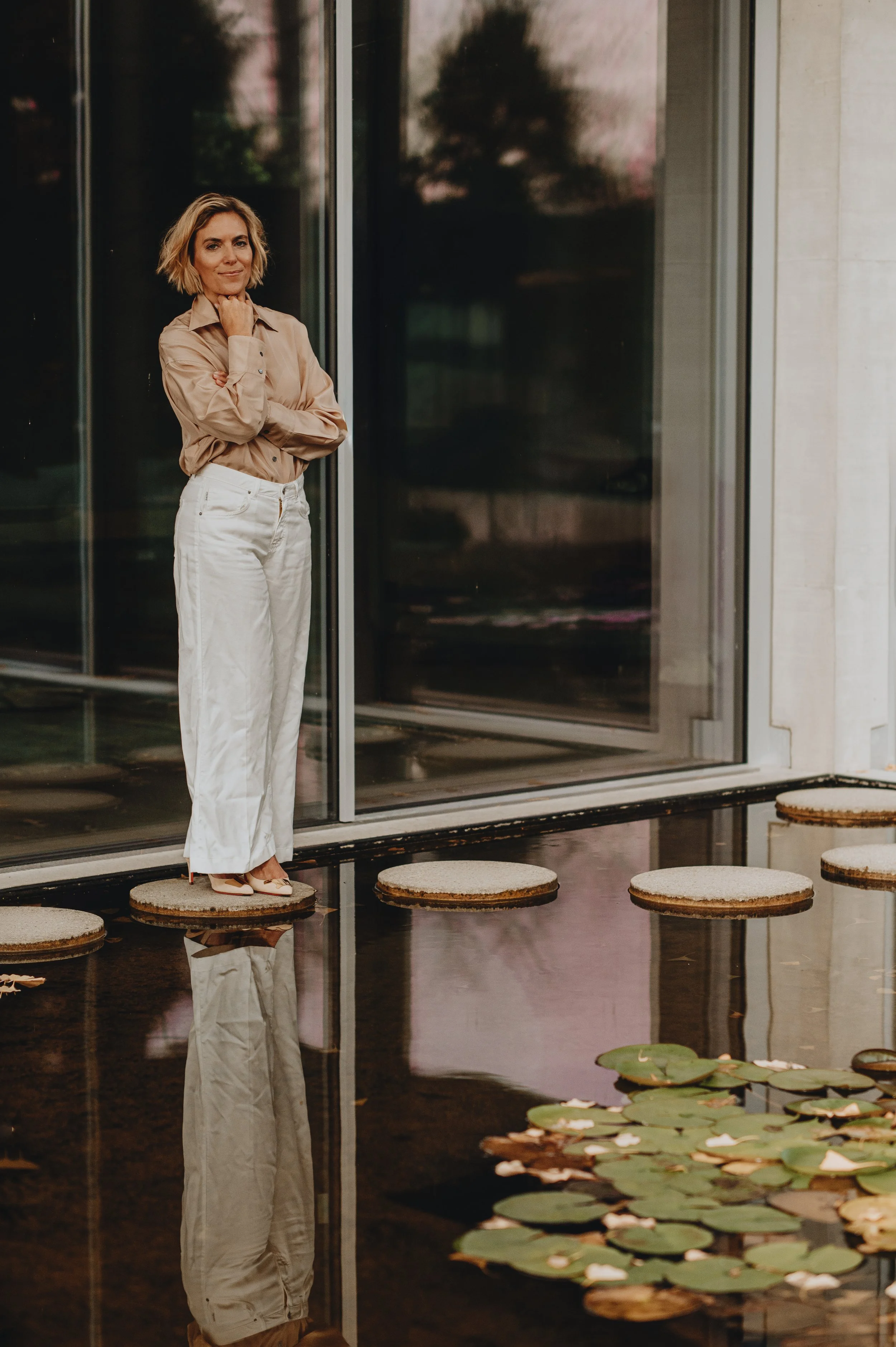 A woman standing on a floating cork step on a pond with lily pads, near a glass building, during sunset.