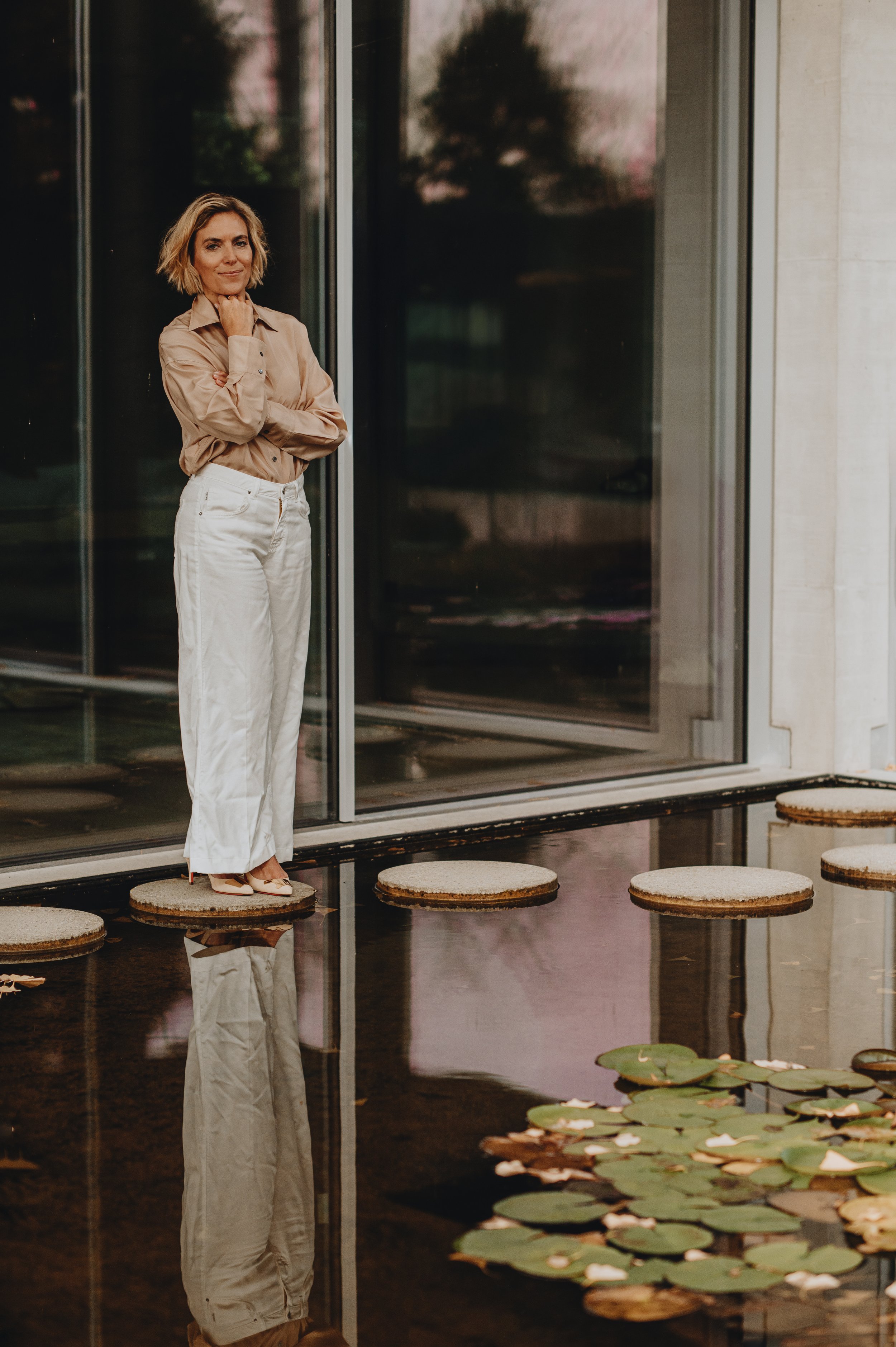 A woman standing on a floating cork step on a pond with lily pads, near a glass building, during sunset.