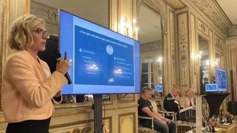 A woman in glasses and a peach blazer holding a microphone, speaking at a presentation in a gilded room. A large screen displays a slide with text, and three people are seated at a table beside her.