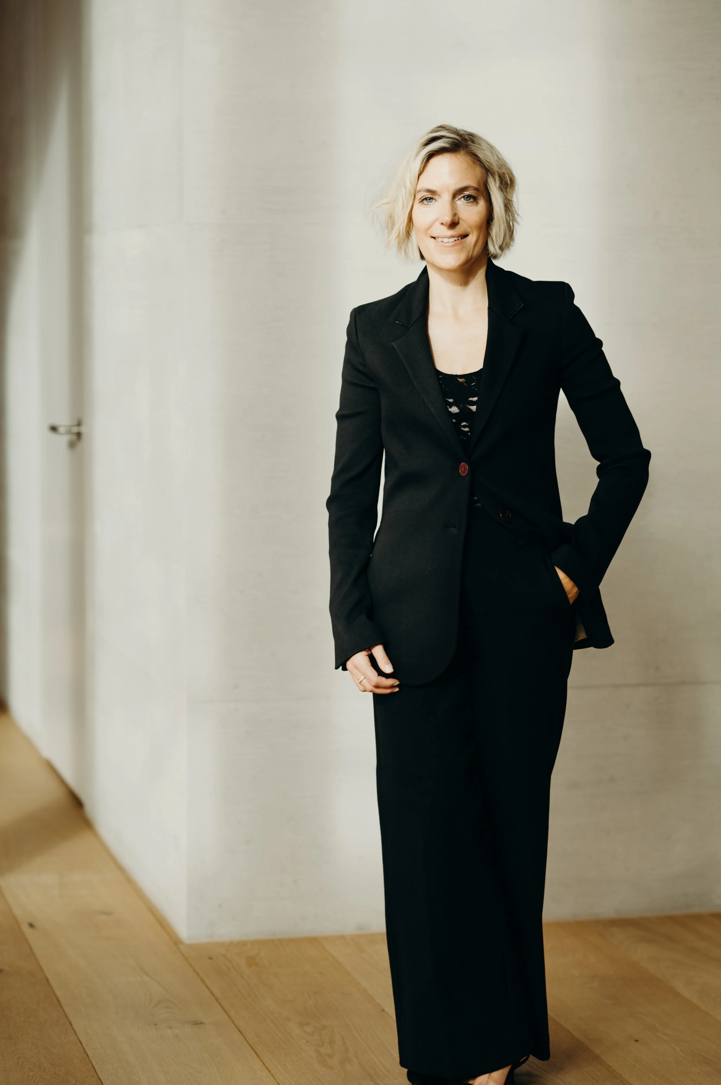 A woman in a black business suit standing in a modern office or home space with a wooden floor and light-colored wall.