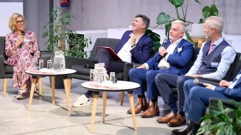 A panel discussion with five people, four men and one woman, in a modern, indoor setting with plants and concrete walls, where the woman is speaking with a microphone and the men are listening, some smiling.