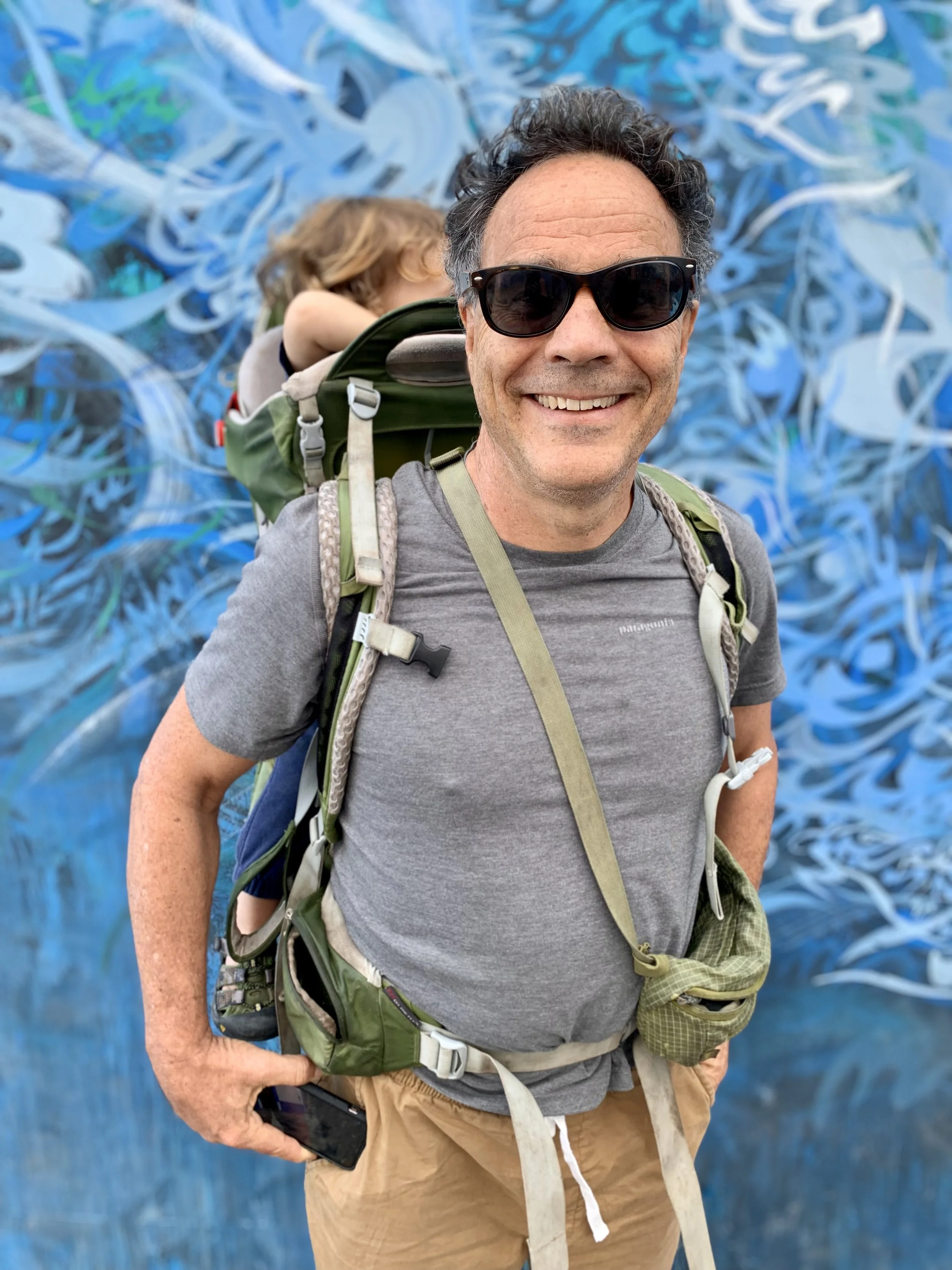 A smiling man with sunglasses wearing a gray t-shirt and beige shorts, standing in front of a blue abstract mural, carrying a green backpack with a child in a child carrier on his back, holding a smartphone in his right hand.