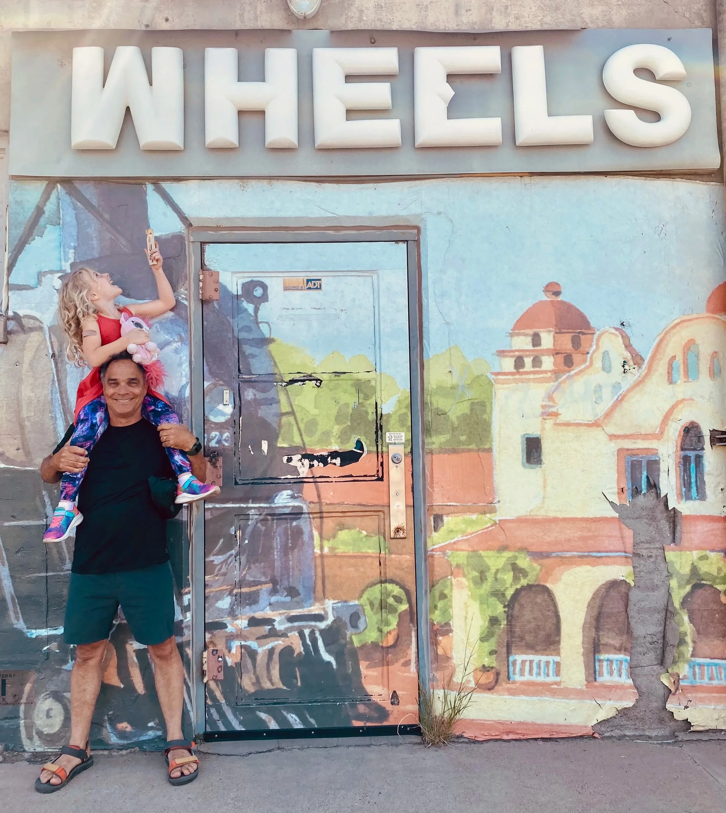 A man is holding a young girl on his shoulders. The girl is taking a selfie with her phone. They are standing in front of a mural with the word 'WHEELS' displayed prominently at the top. The mural depicts buildings and a scenic background.