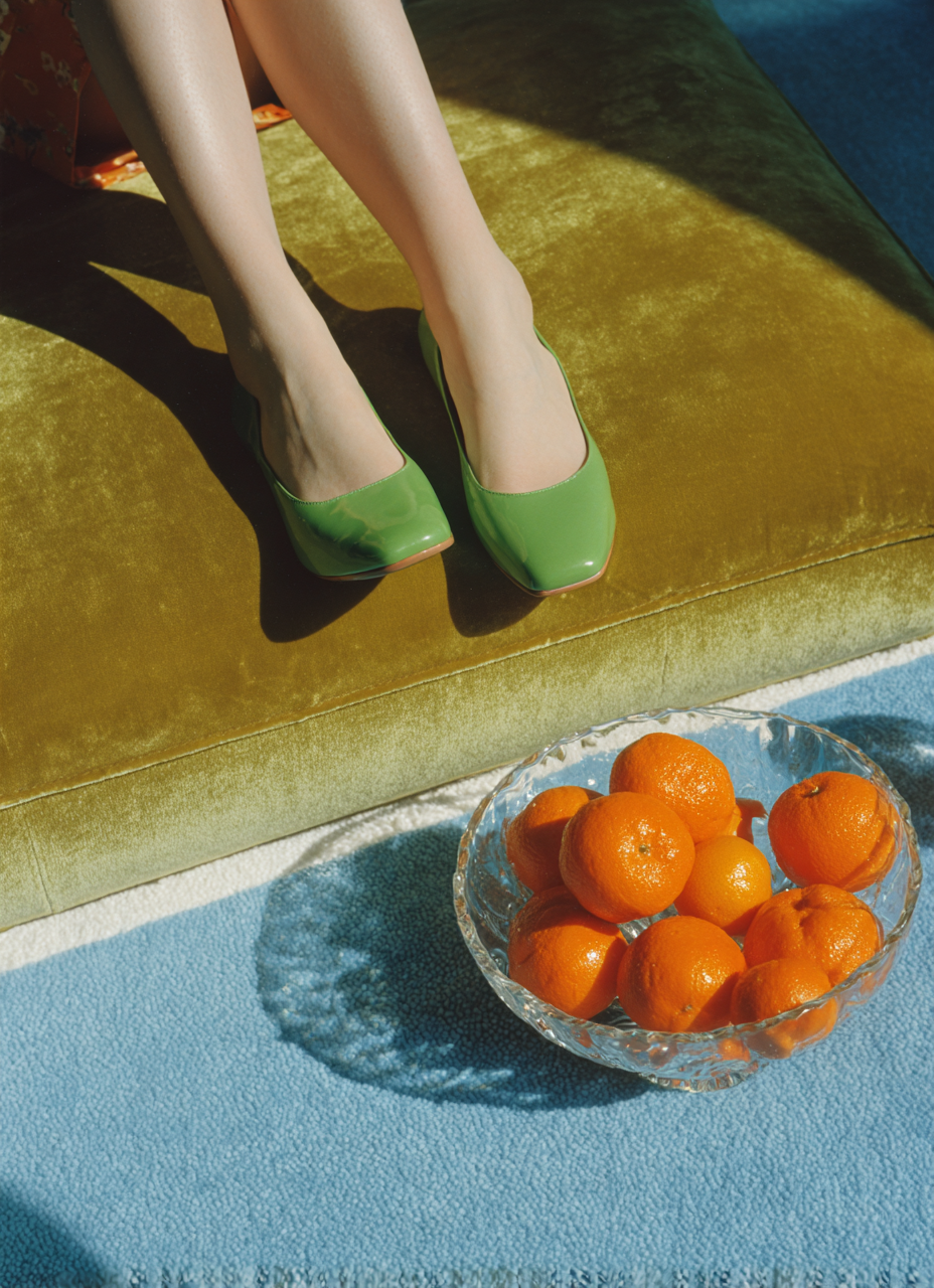 Piedi di una persona con calze e scarpe verdi su un divano verde, con un piatto di arance sul pavimento vicino.