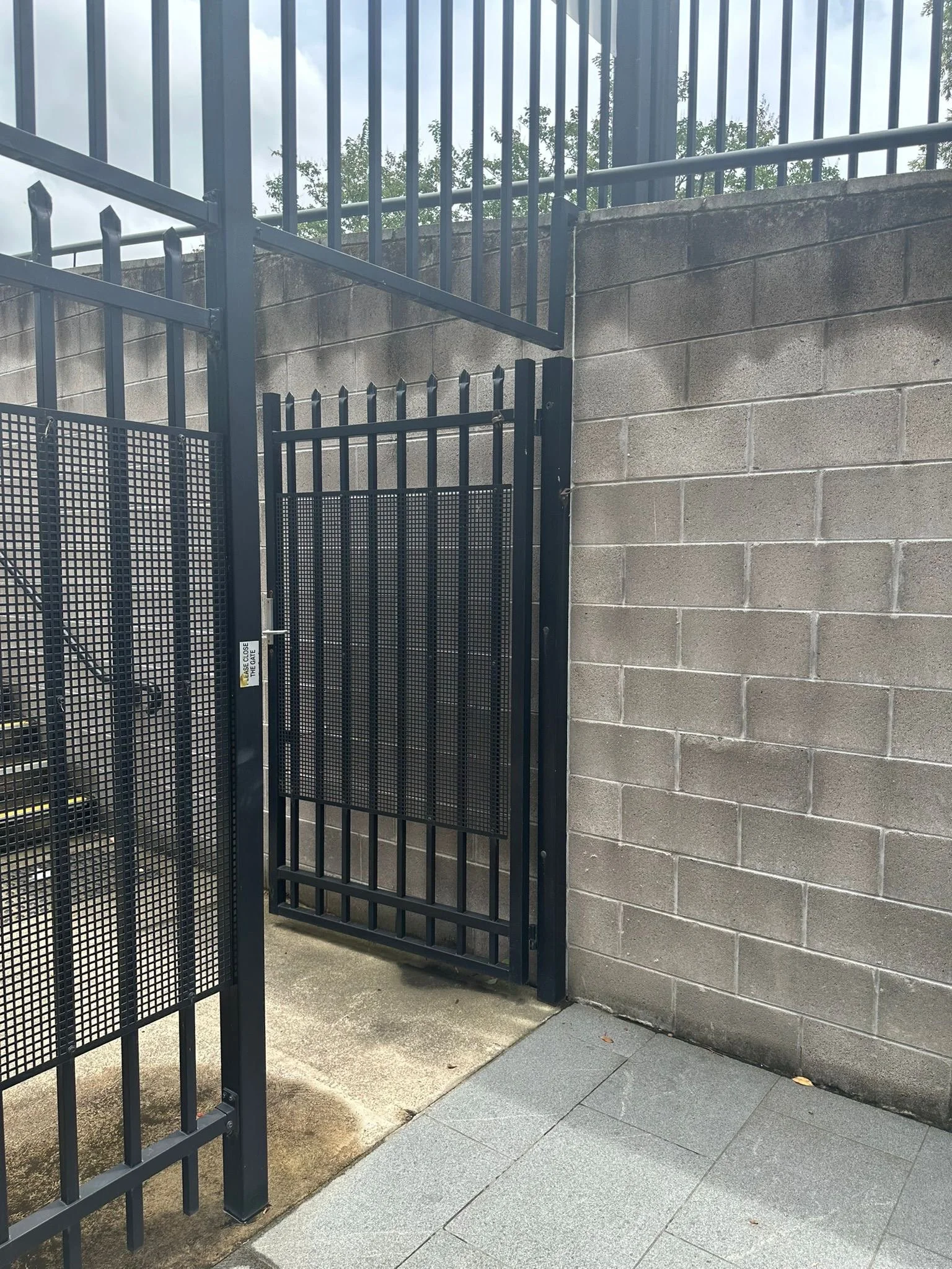 Image of a black metal gate with a mesh panel, attached to a gray concrete block wall, with a concrete and tiled ground area.