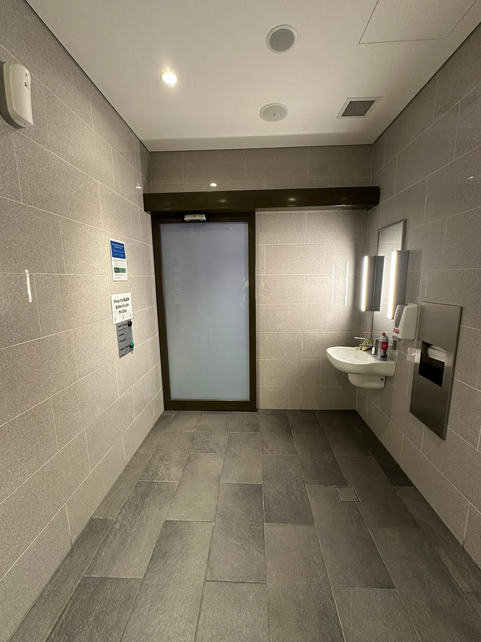 Empty public restroom with a small sink, soap dispenser, paper towel dispenser, mirror, and frosted glass door.
