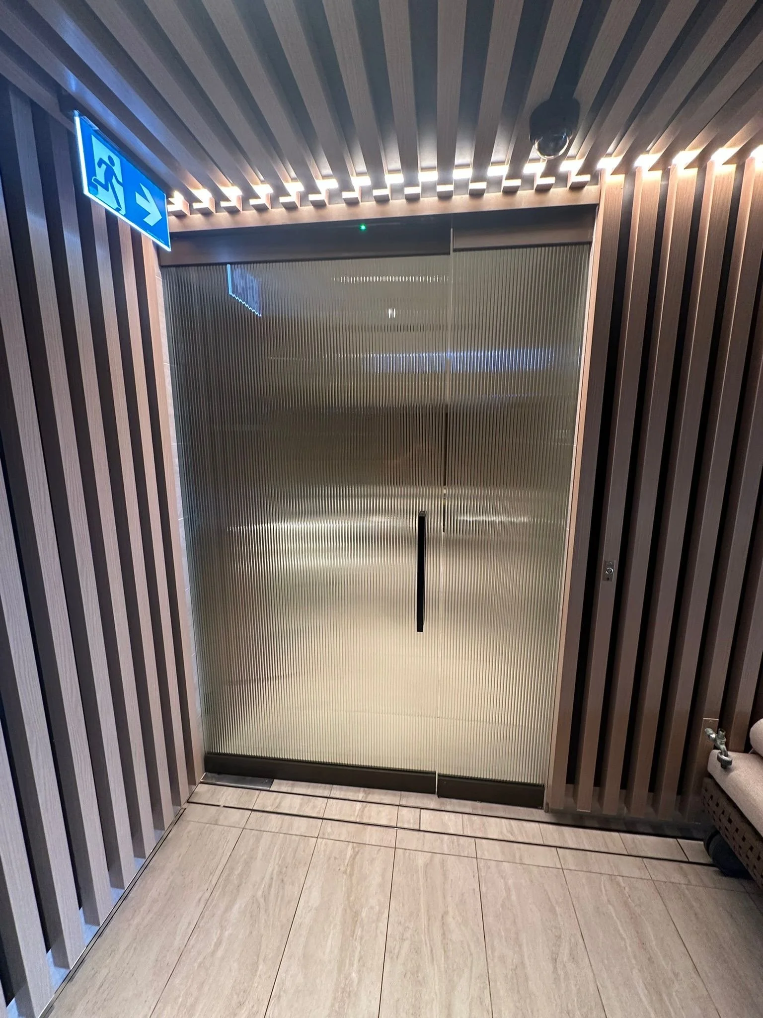 A stainless steel sliding door with vertical ribbing, surrounded by wood-paneled walls and a beige tiled floor, with an emergency exit sign above.