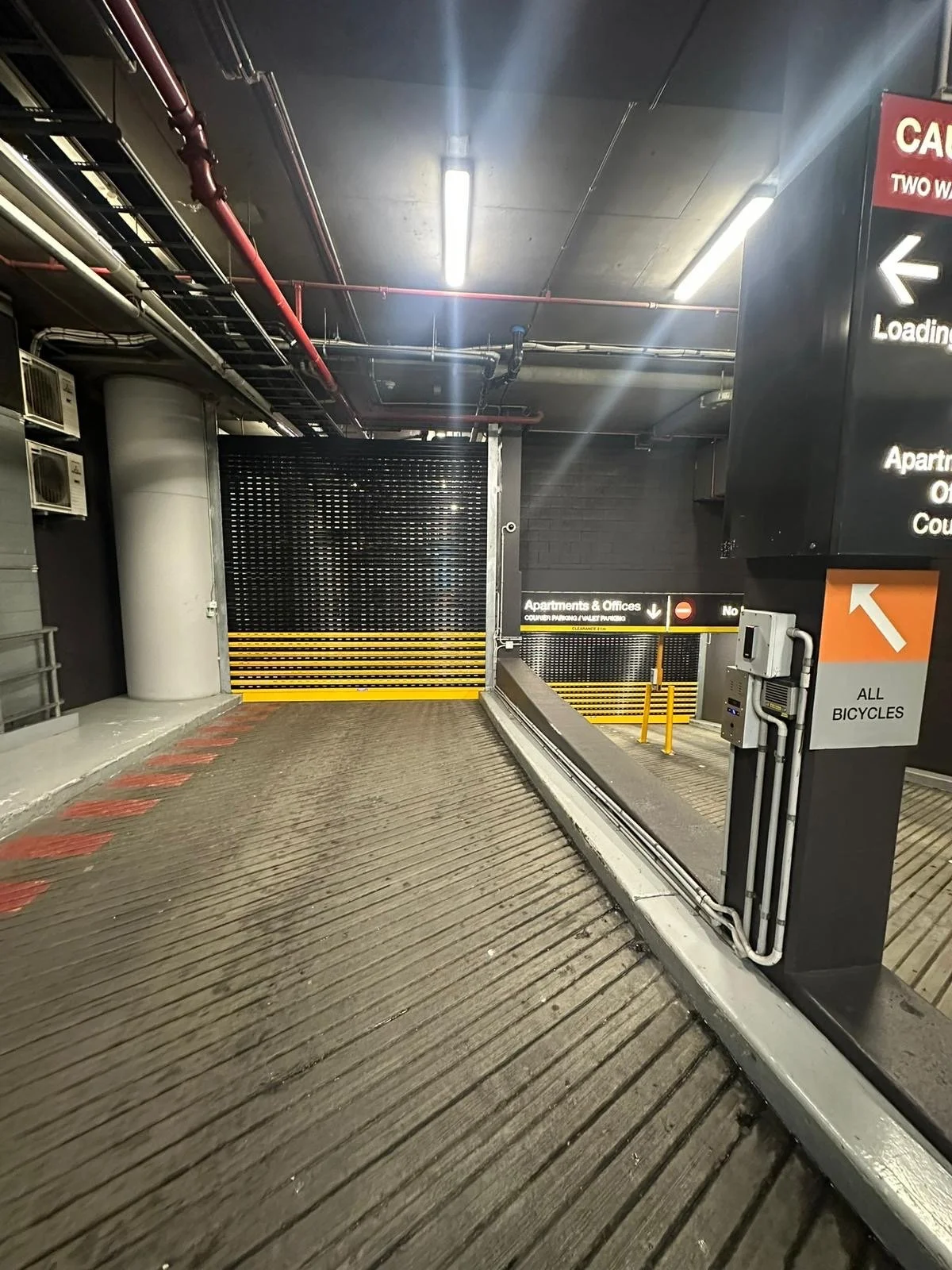 Entrance of a parking garage ramp with a closed black gate, warning signs, and directional signage for bicycles and buildings.