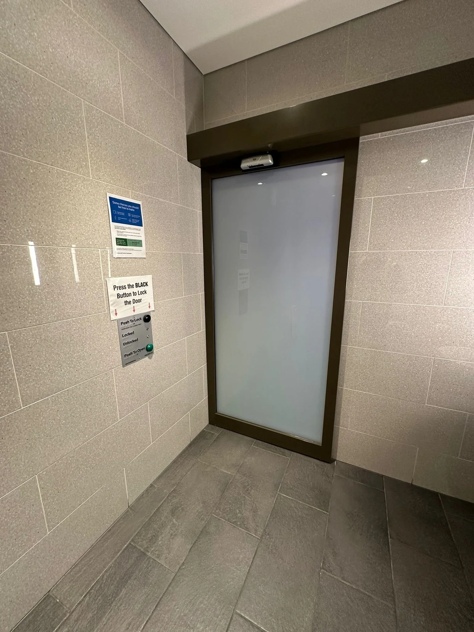 A glass door with a metal frame in a tiled wall, equipped with an intercom or lock system with instructions to press the black button to lock the door, and push to open.