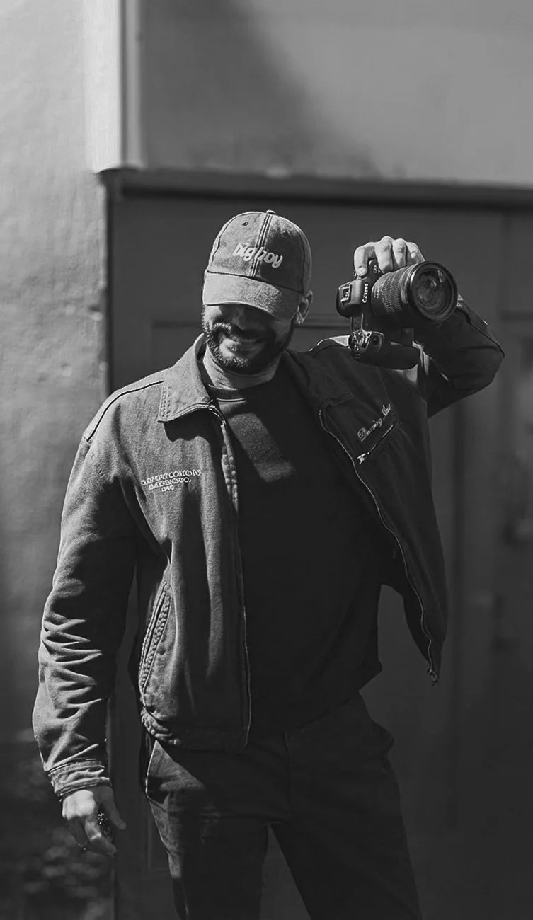 Le photographe souriant portant une casquette et un blouson, tenant un appareil photo à la main.