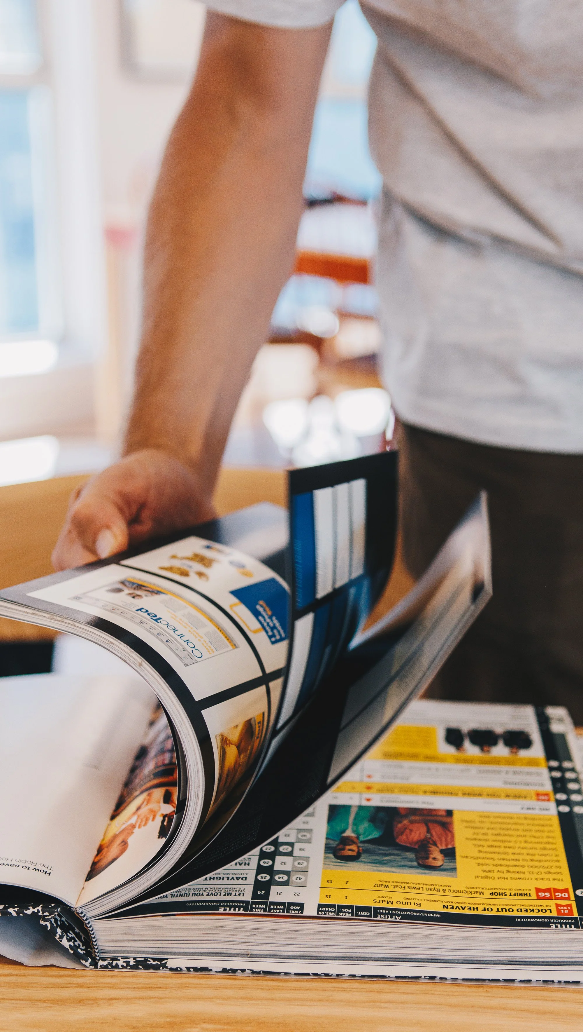 A person flipping through a brochure or magazine on a table, with another magazine or newspaper underneath.