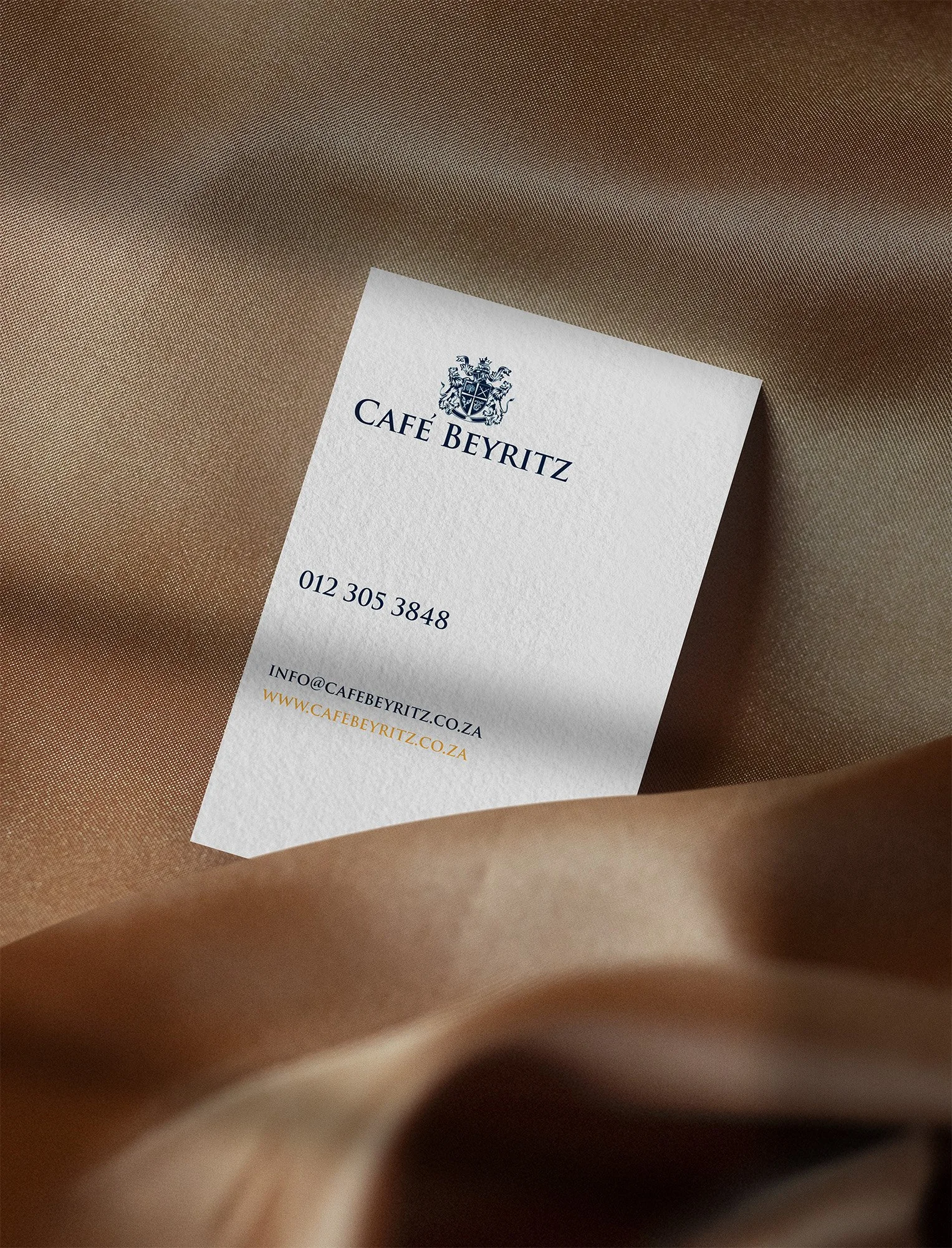 A business card for Cafe Beyritz with contact information, placed on a beige satin fabric background.