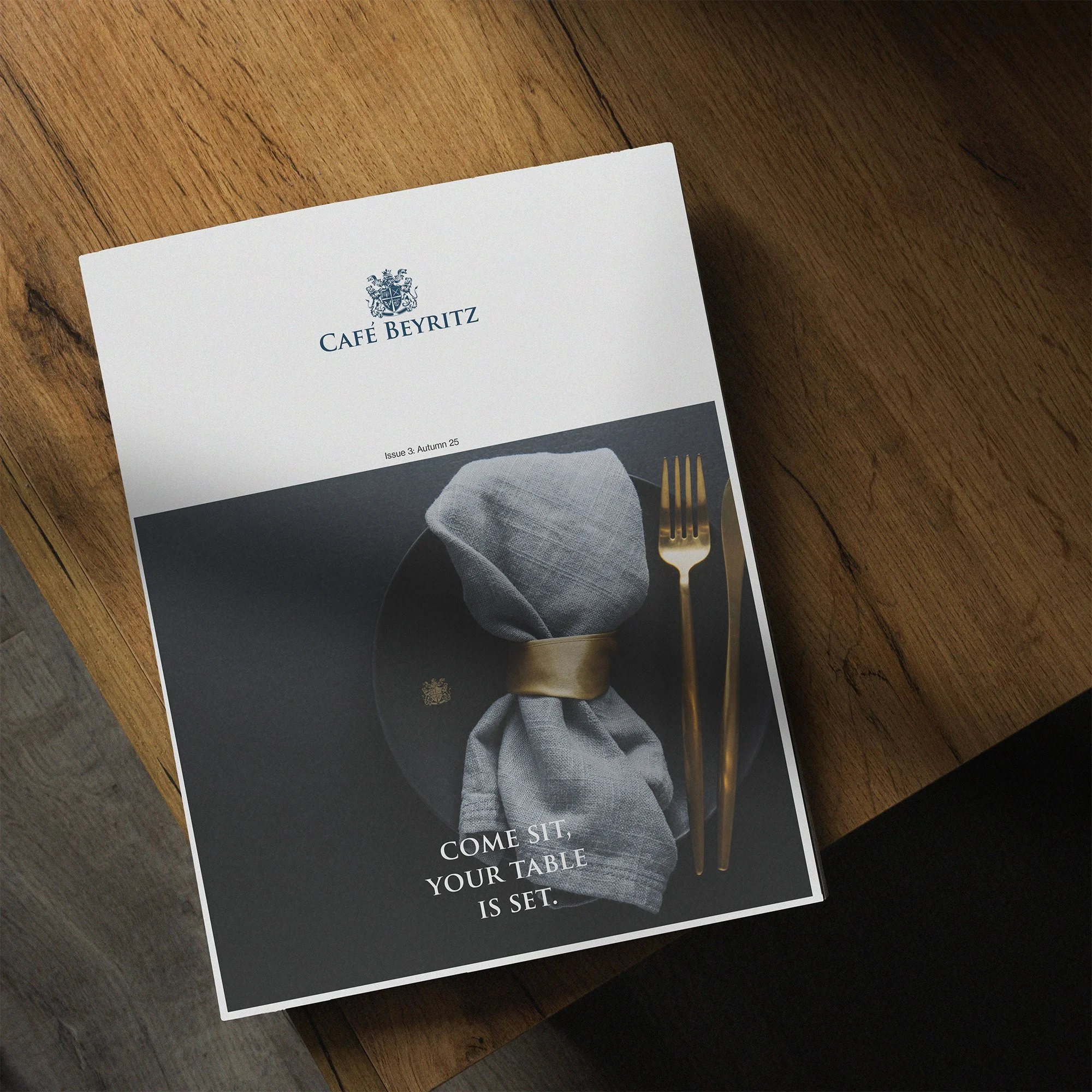 A printed menu from Café Beyritz resting on a wooden table with a black and white cover, featuring a wrapped napkin, a gold fork, and knife, and the text 'Come sit, your table is set'.