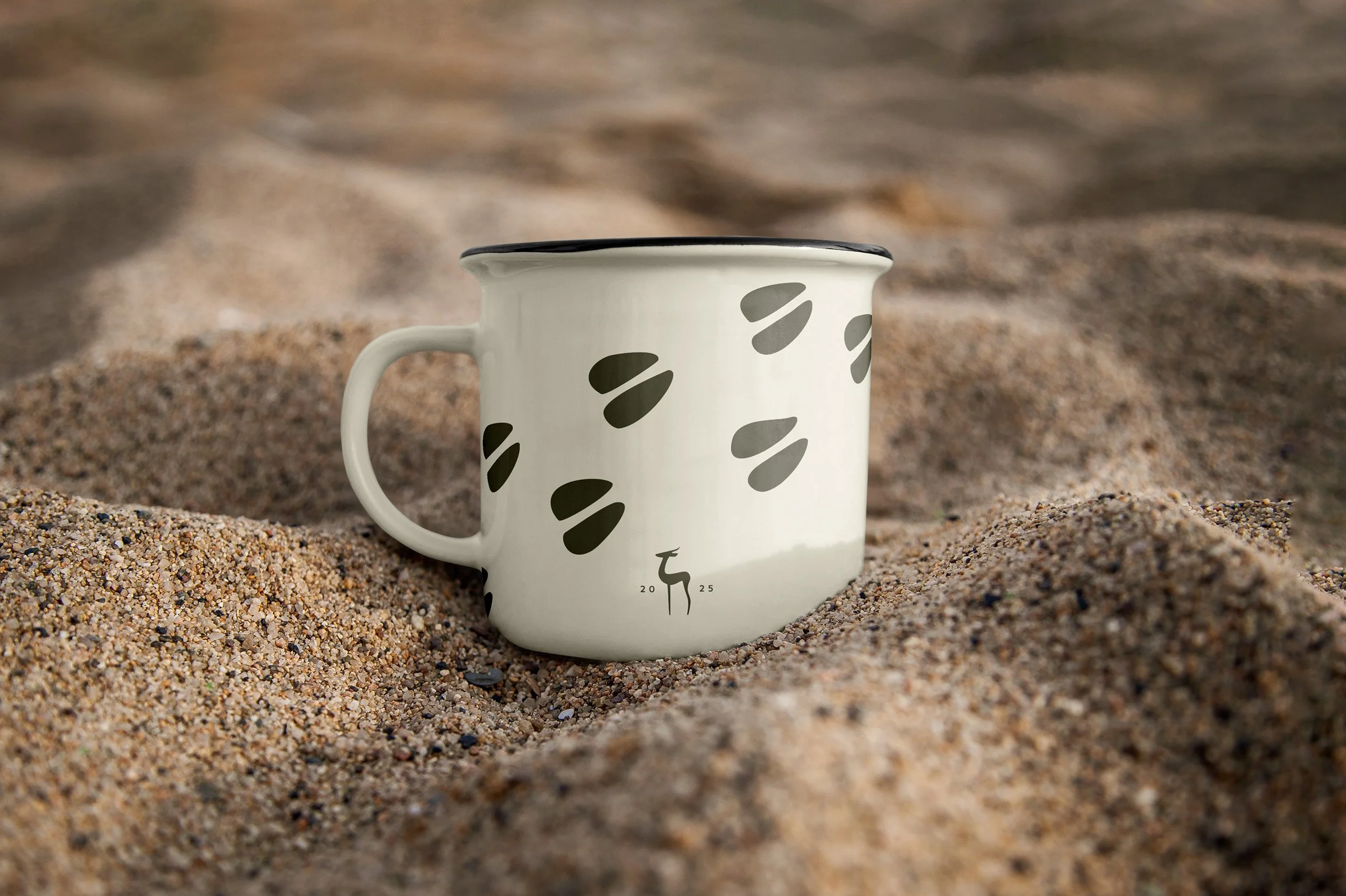 A white enamel mug with black abstract spots positioned on sandy ground with blurred sand in the background.