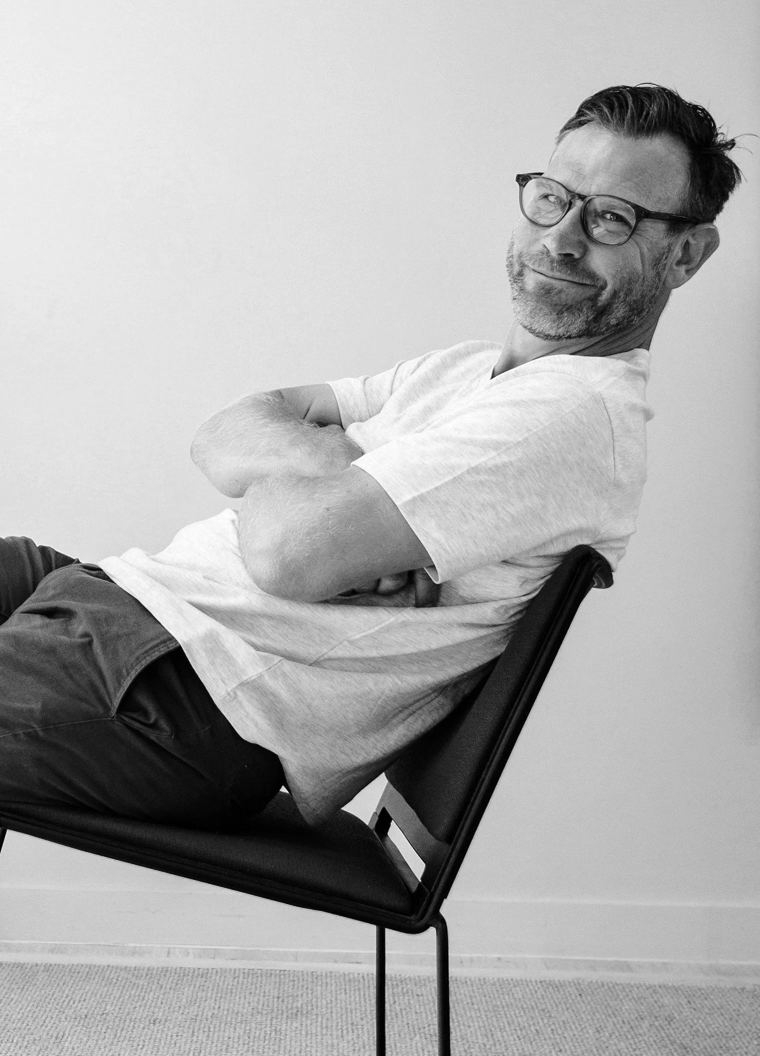 A middle-aged man with glasses and facial hair sitting in a chair with his arms crossed, smiling slightly, in front of a plain wall.