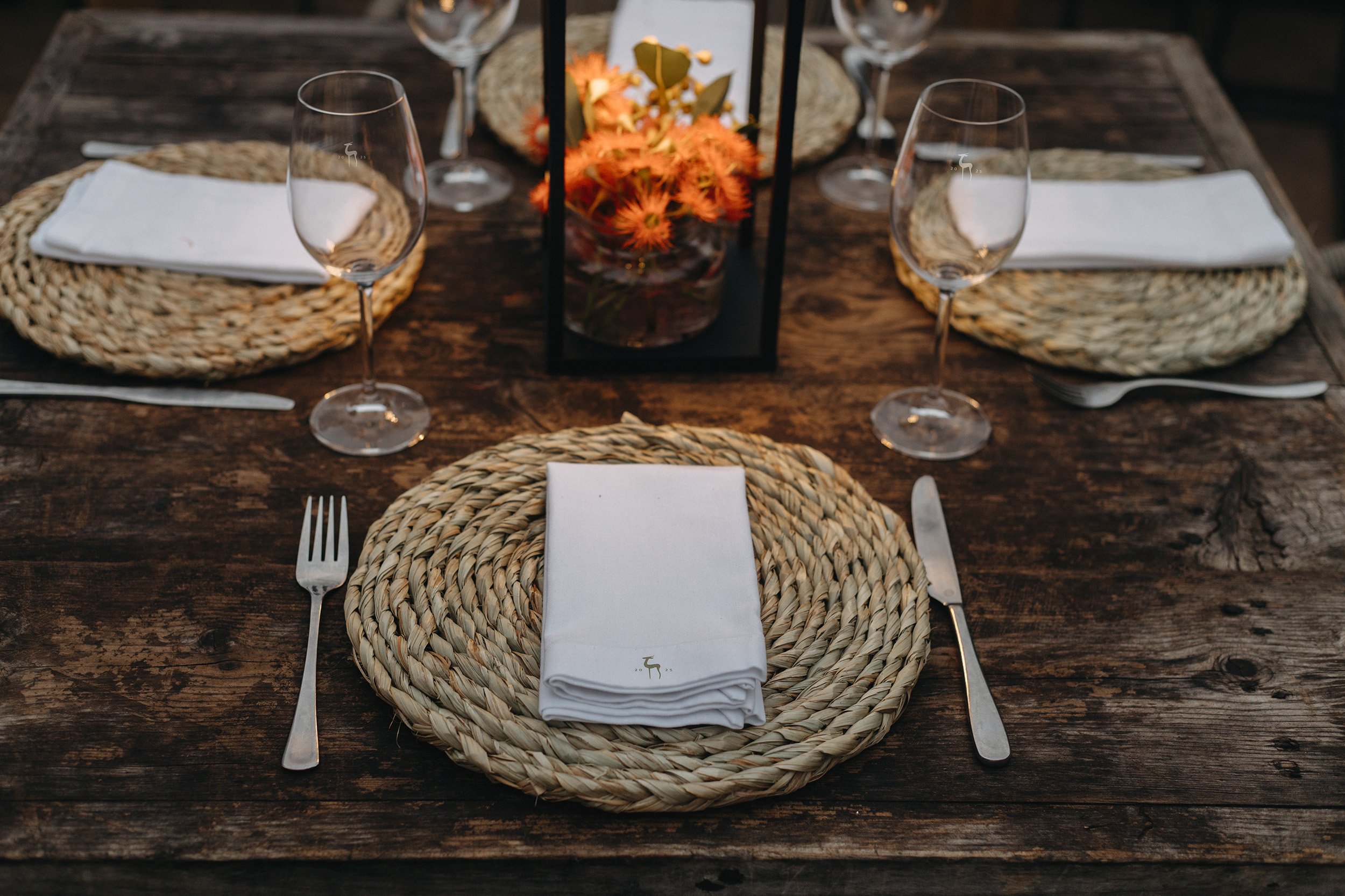 A rustic wooden dining table set with four woven placemats, white napkins, wine glasses, and a centerpiece of orange flowers in a black frame.